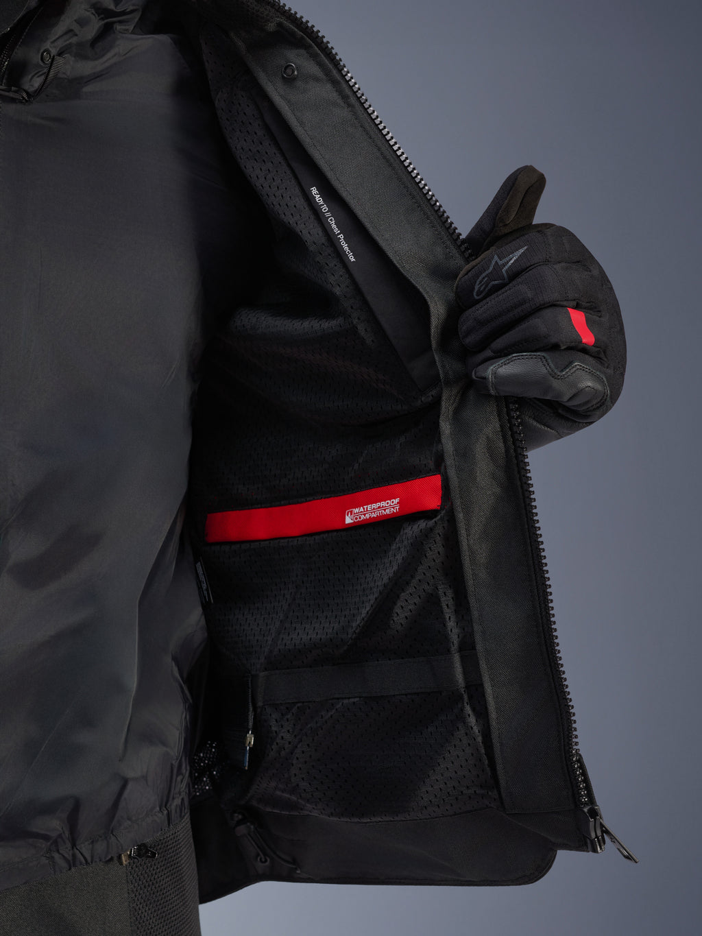 Indvendigt detaljebillede af Alpinestars Andes Air Drystar®-jakken, der fremhæver det åndbare sorte mesh-for, det røde vandtætte rum og lommen til brystbeskytteren, vist holdt åben af en sort motorcykelhandske.