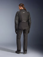 Kvinde set bagfra iført Alpinestars Stella Andes V4 Drystar® jakke, sort, matchende motorcykelbukser, hvidt Astar-logo på ryggen, forlænget rygstykke, komplet adventure-touring køreudstyr inklusiv sorte handsker og støvler