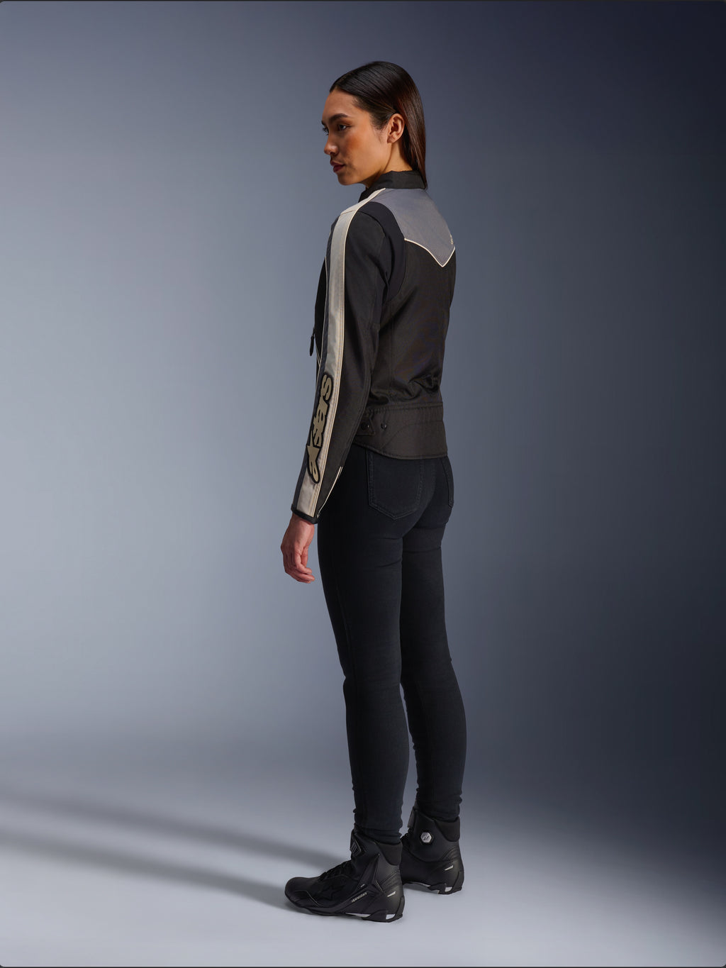 Kvindelig model set fra siden og bagfra iført Alpinestars Stella T-Dyno WR-jakken i Black Dark Gray Ecru, med vintage-inspireret design med ecrufarvede striber på ærmerne og et gråt rygpanel, båret med sorte bukser og sorte køresko.