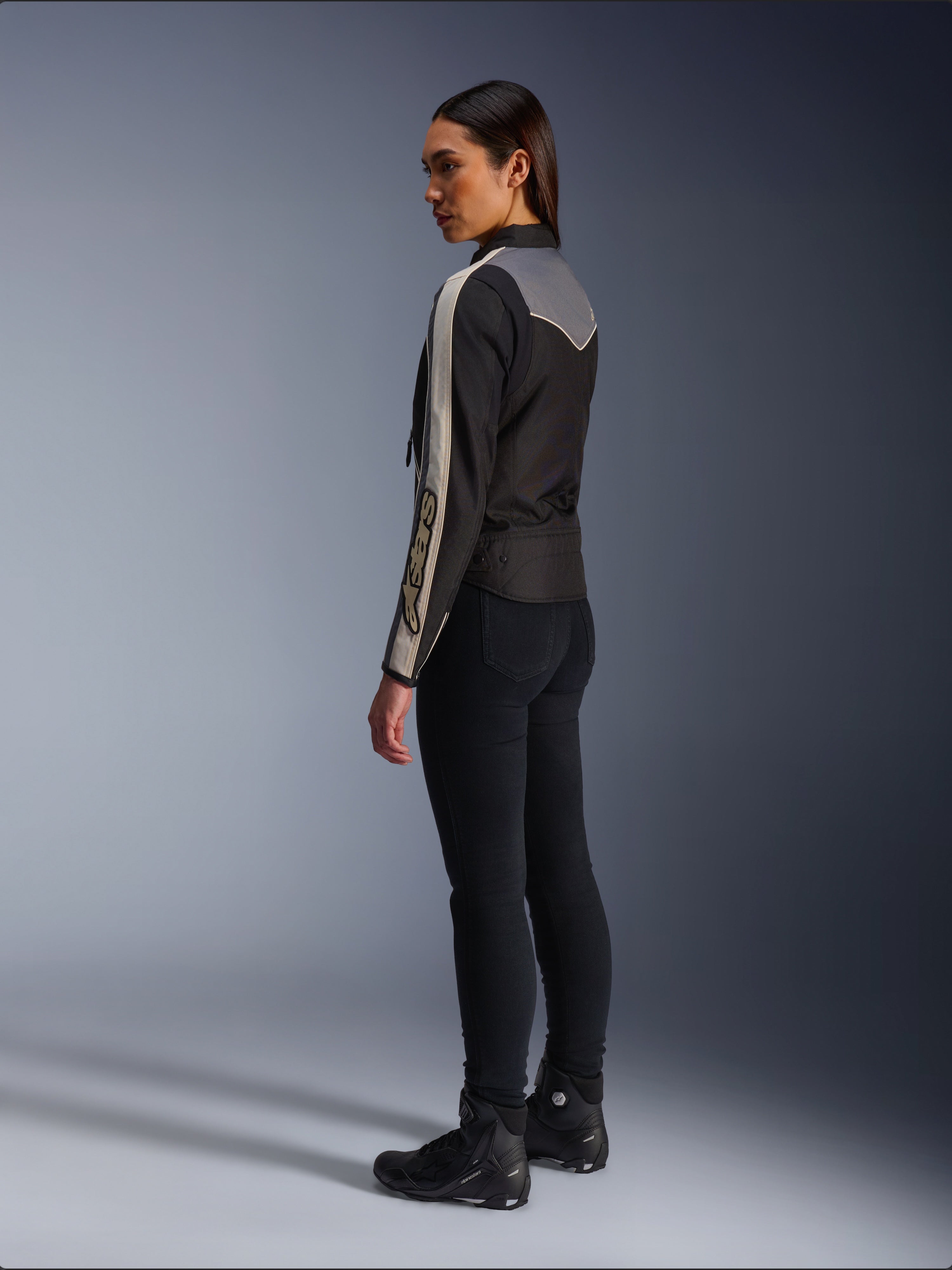 Kvindelig model set fra siden og bagfra iført Alpinestars Stella T-Dyno WR-jakken i Black Dark Gray Ecru, med vintage-inspireret design med ecrufarvede striber på ærmerne og et gråt rygpanel, båret med sorte bukser og sorte køresko.