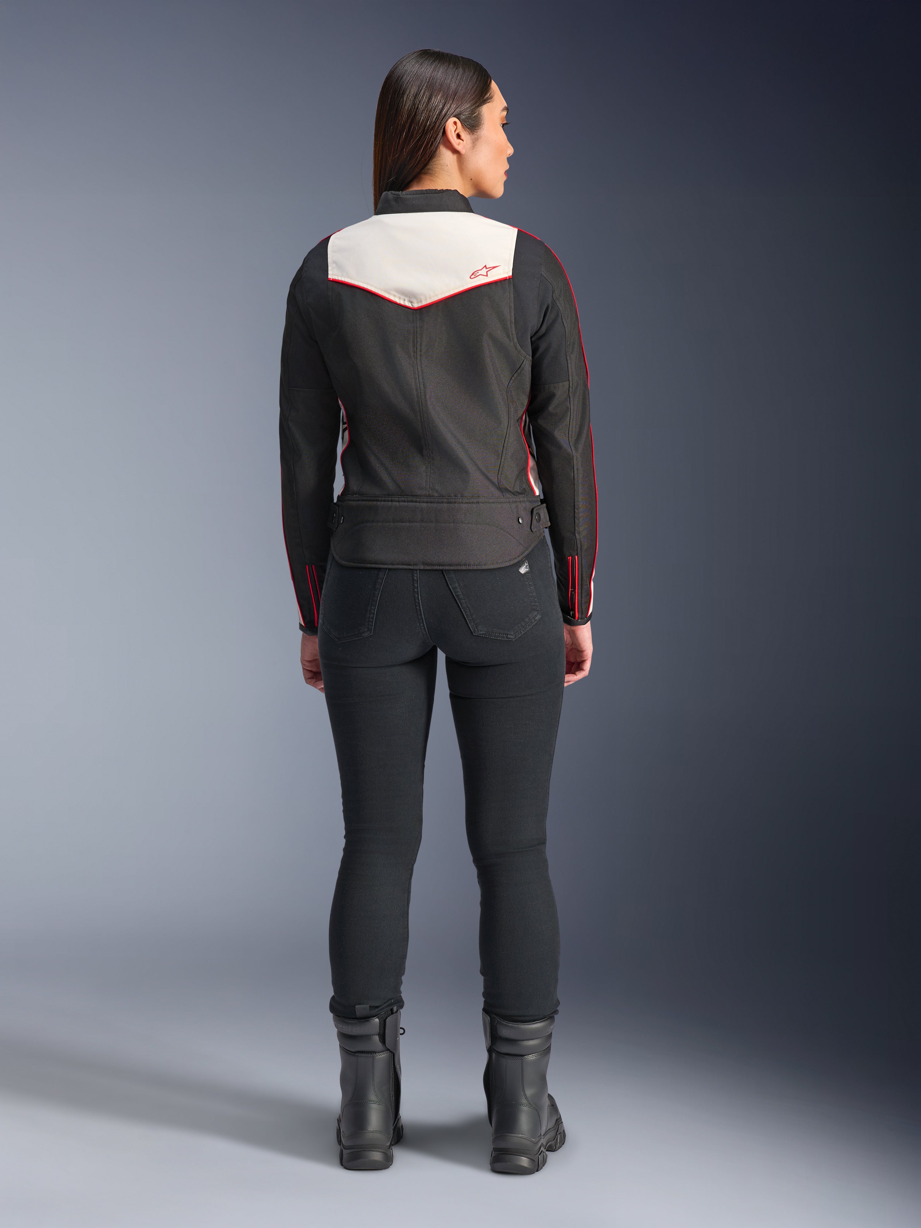 Bagfra set af en kvindelig model iført Alpinestars Stella T-Dyno WR-jakken i Black Ecru Bright Red, med et fremtrædende cremefarvet rygpanel og røde logo-detaljer, stylet med sorte bukser og støvler, en vintage-inspireret tekstil-motorcykeljakke.