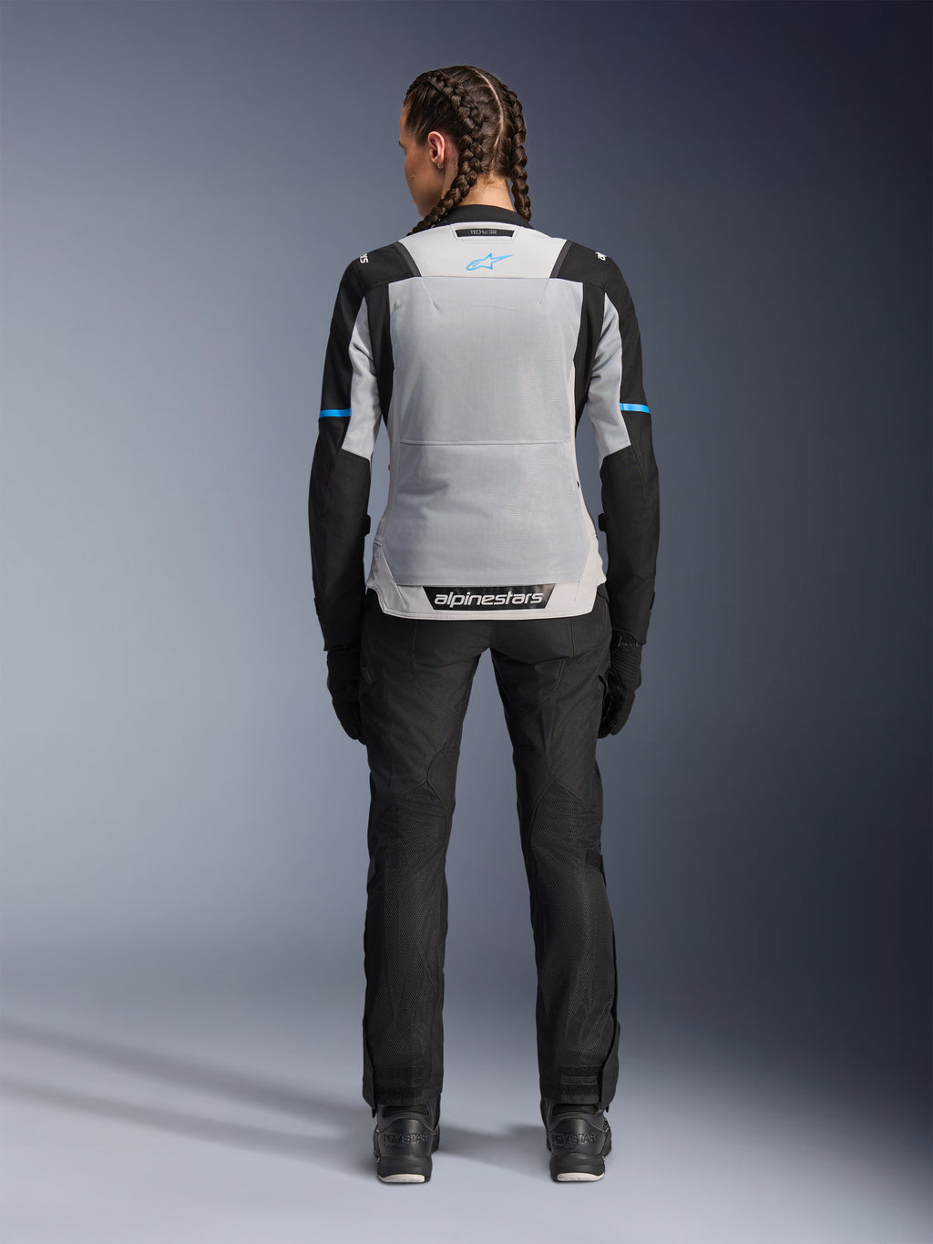 Alpinestars Stella ST-2 Air jakke, Ice Gray Black, set bagfra på en kvindelig model, med et stort lysegråt mesh-rygpanel, sorte ærmer med blå stribedetaljer, Alpinestars-branding på den nederste del af ryggen, kombineret med sorte tekniske kørebukser og støvler, sommer-motorcykel-touringudstyr