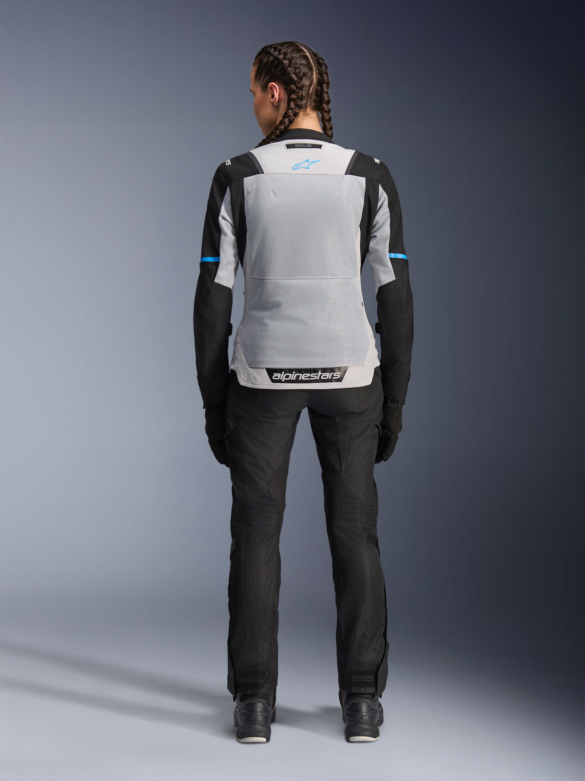 Alpinestars Stella ST-2 Air jakke, Ice Gray Black, set bagfra på en kvindelig model, med et stort lysegråt mesh-rygpanel, sorte ærmer med blå stribedetaljer, Alpinestars-branding på den nederste del af ryggen, kombineret med sorte tekniske kørebukser og støvler, sommer-motorcykel-touringudstyr