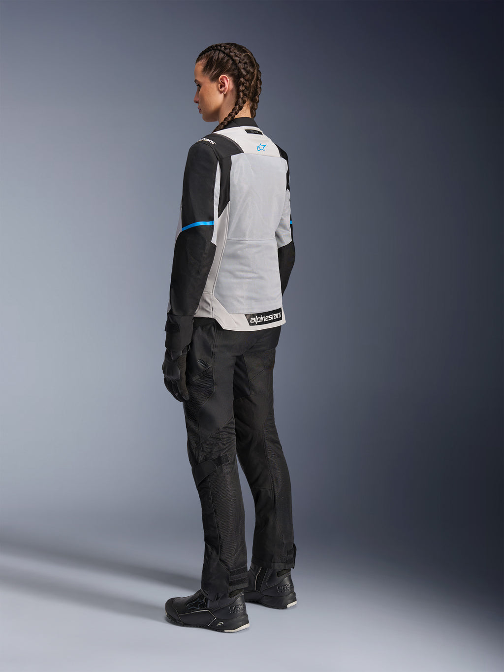 Kvindelig motorcyklist iført Alpinestars Stella ST-2 Air-jakke i Ice Gray og sort, set bagfra i trekvart profil, der fremhæver det store lysegrå mesh-rygpanel, sorte ærmer med en blå accentstribe og et blåt Alpinestars-logo, kombineret med sorte motorcykelbukser, handsker og støvler for et komplet adventure touring-outfit.
