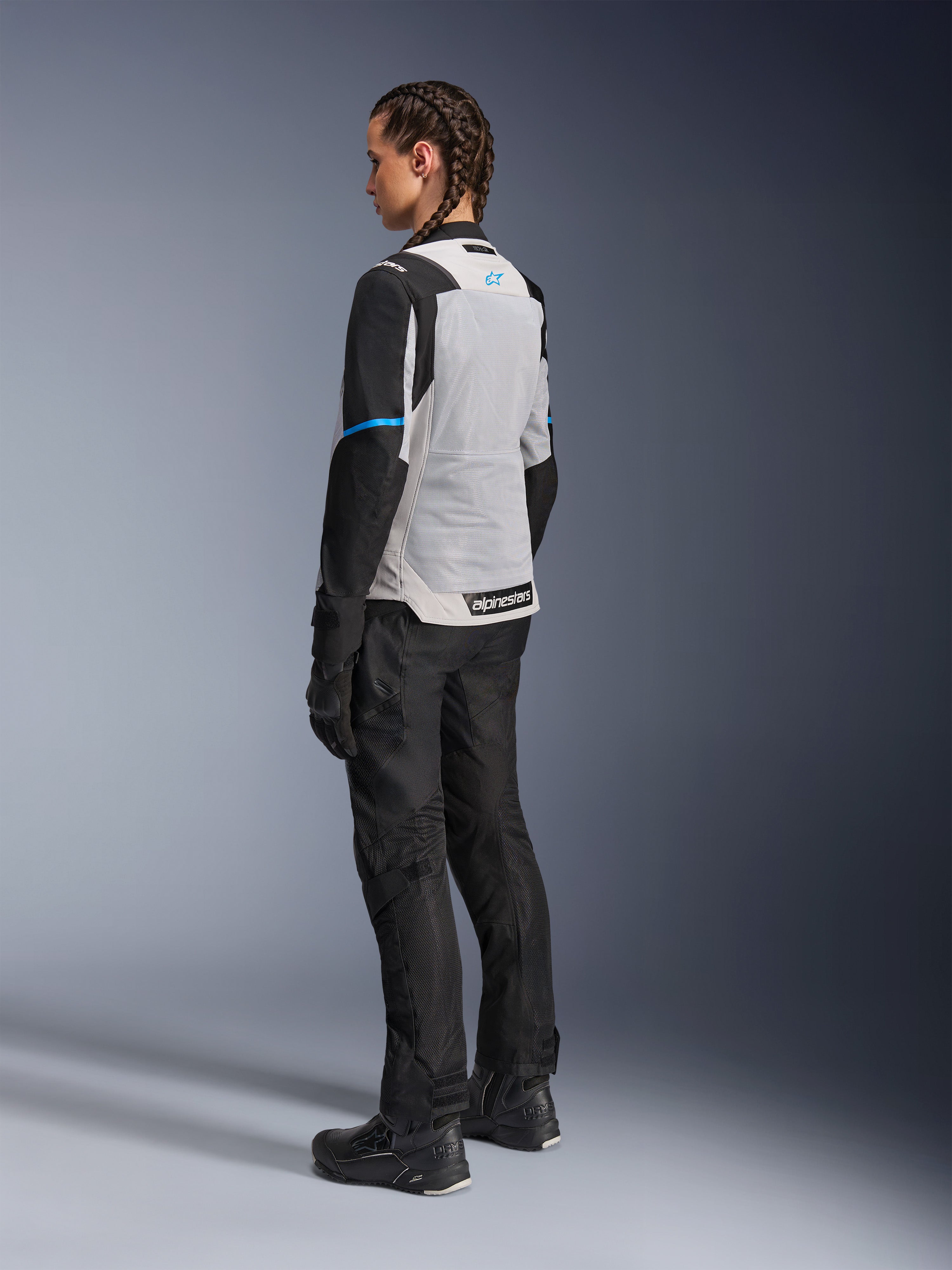 Kvindelig motorcyklist iført Alpinestars Stella ST-2 Air-jakke i Ice Gray og sort, set bagfra i trekvart profil, der fremhæver det store lysegrå mesh-rygpanel, sorte ærmer med en blå accentstribe og et blåt Alpinestars-logo, kombineret med sorte motorcykelbukser, handsker og støvler for et komplet adventure touring-outfit.