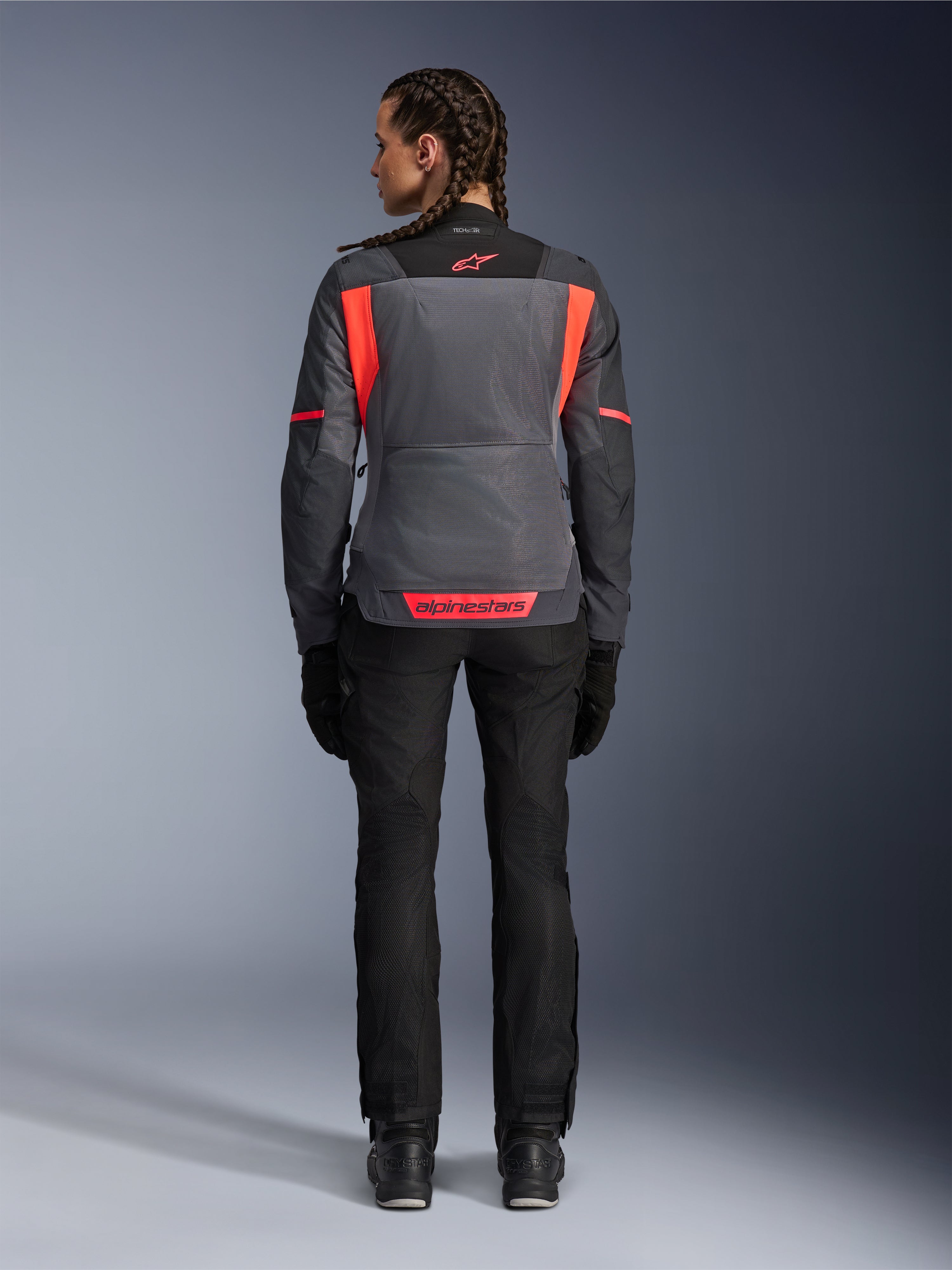 Rygbillede af en model iført Alpinestars Woman Stella St-2 Air Jacket i Deep Gray og Dark Gray med Fluo Coral-detaljer, med store mesh-ventilationspaneler på ryggen og reflekterende logodetaljer, kombineret med sorte motorcykelbukser og -handsker.