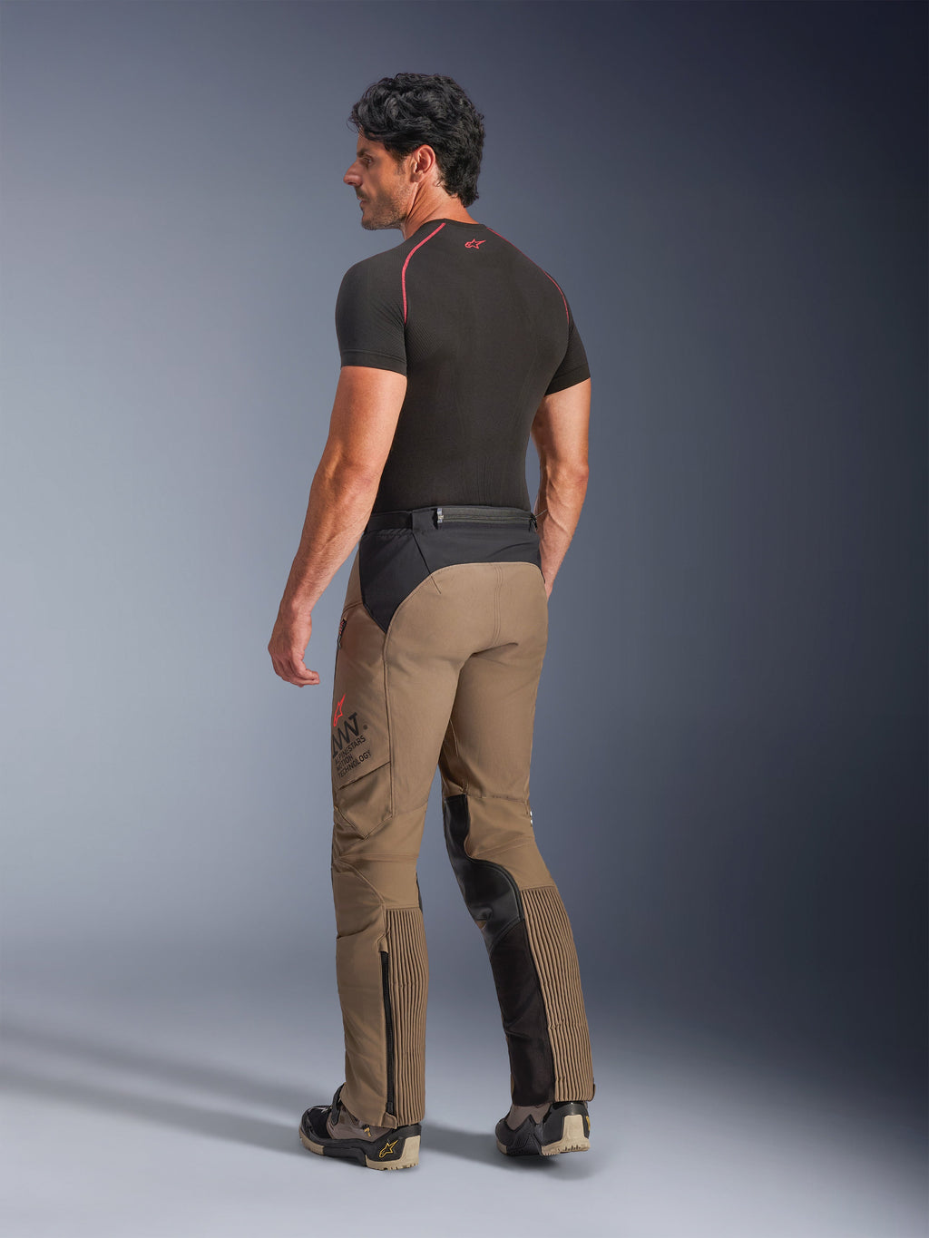 Alpinestars AMT-8 Stretch Drystar® XF bukser i Walnut Black, adventure-touring motorcykelbukser vist skråt bagfra, med nøddebrunt tekstil og sorte paneler på sædet og indersiden af benene, harmonika-strækpaneler på læggene og AMT-branding på siden, båret med et sort baselayer.