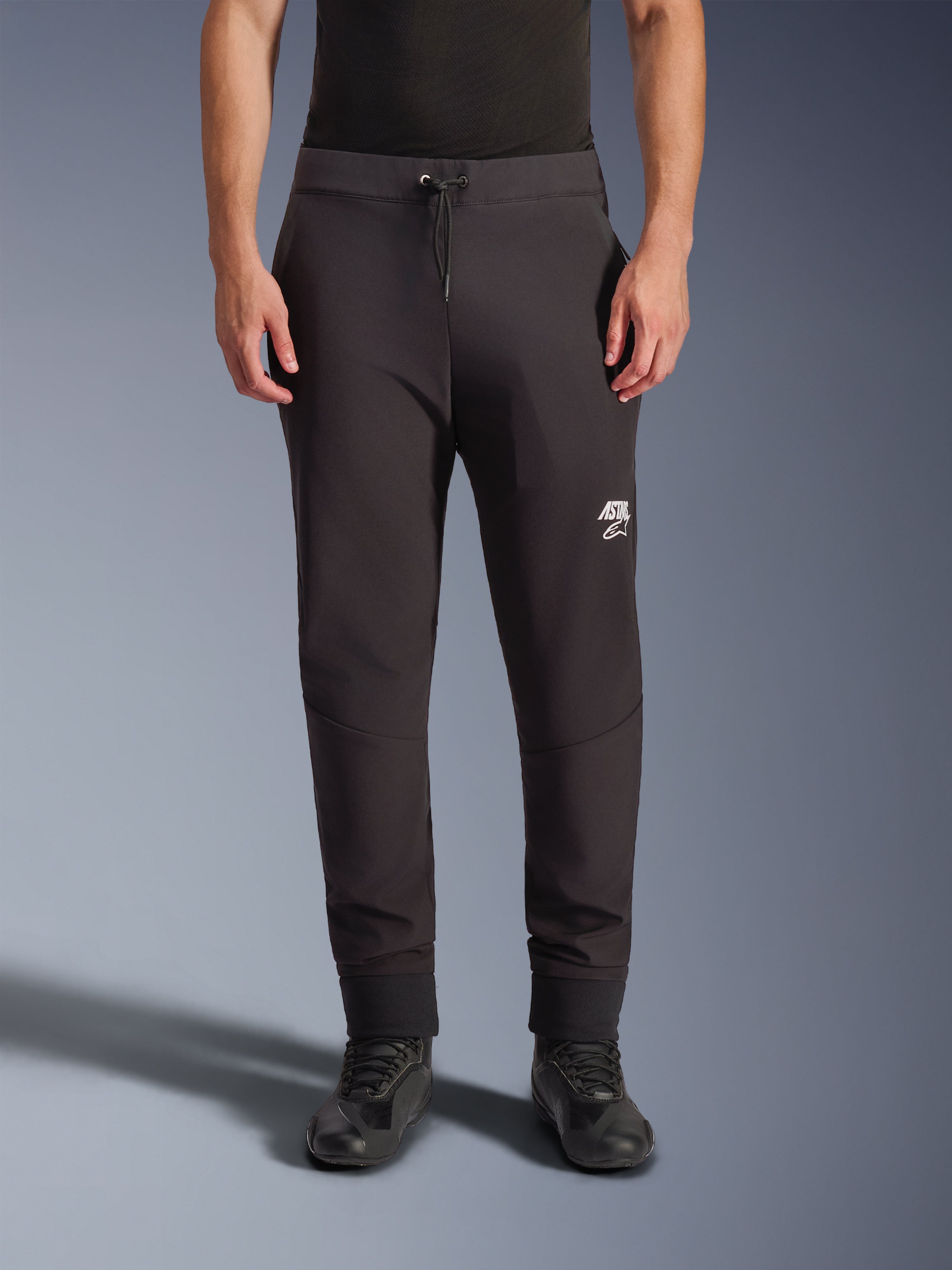 Alpinestars Aeroshell bukser, sorte softshell motorcykelbukser, atletisk pasform i jogger-stil med snøre i taljen og elastiske opslag, hvidt Astars-logo, kombineret med sorte køresko, moderne crossover-gear til vej- og fritidsbrug