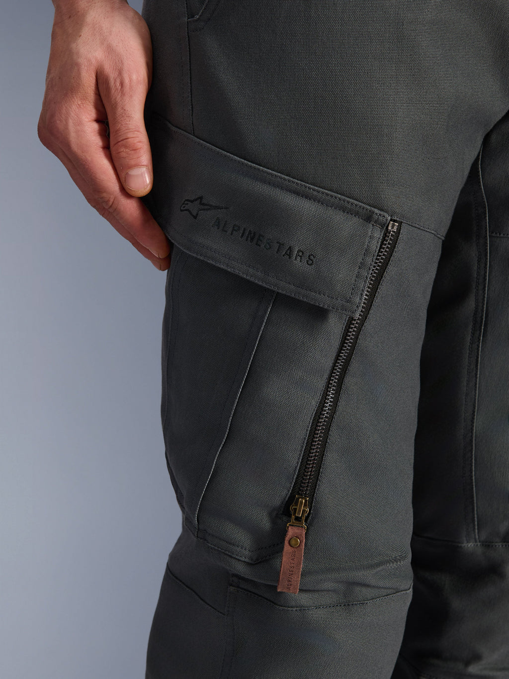 Alpinestars Borrego Drystar® bukser, antracitgrå, nærbillede af cargolomme på låret med broderet branding, tekstureret voksbehandlet lærredsmateriale, lodret ventilationslynlås med trækstrop i brunt læder