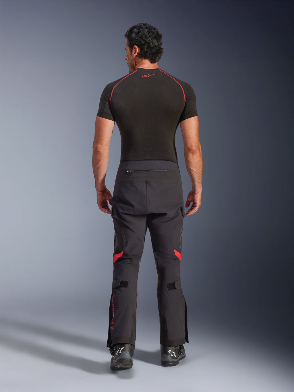 Alpinestars Halo Pro Drystar® XF laminerede bukser, adventure-motorcykelbukser, sort Dark Shadow med røde detaljer, set bagfra kombineret med et sort kortærmet basislag og sorte adventure-støvler, der viser den holdbare 3-lags laminerede konstruktion og ergonomiske pasform.