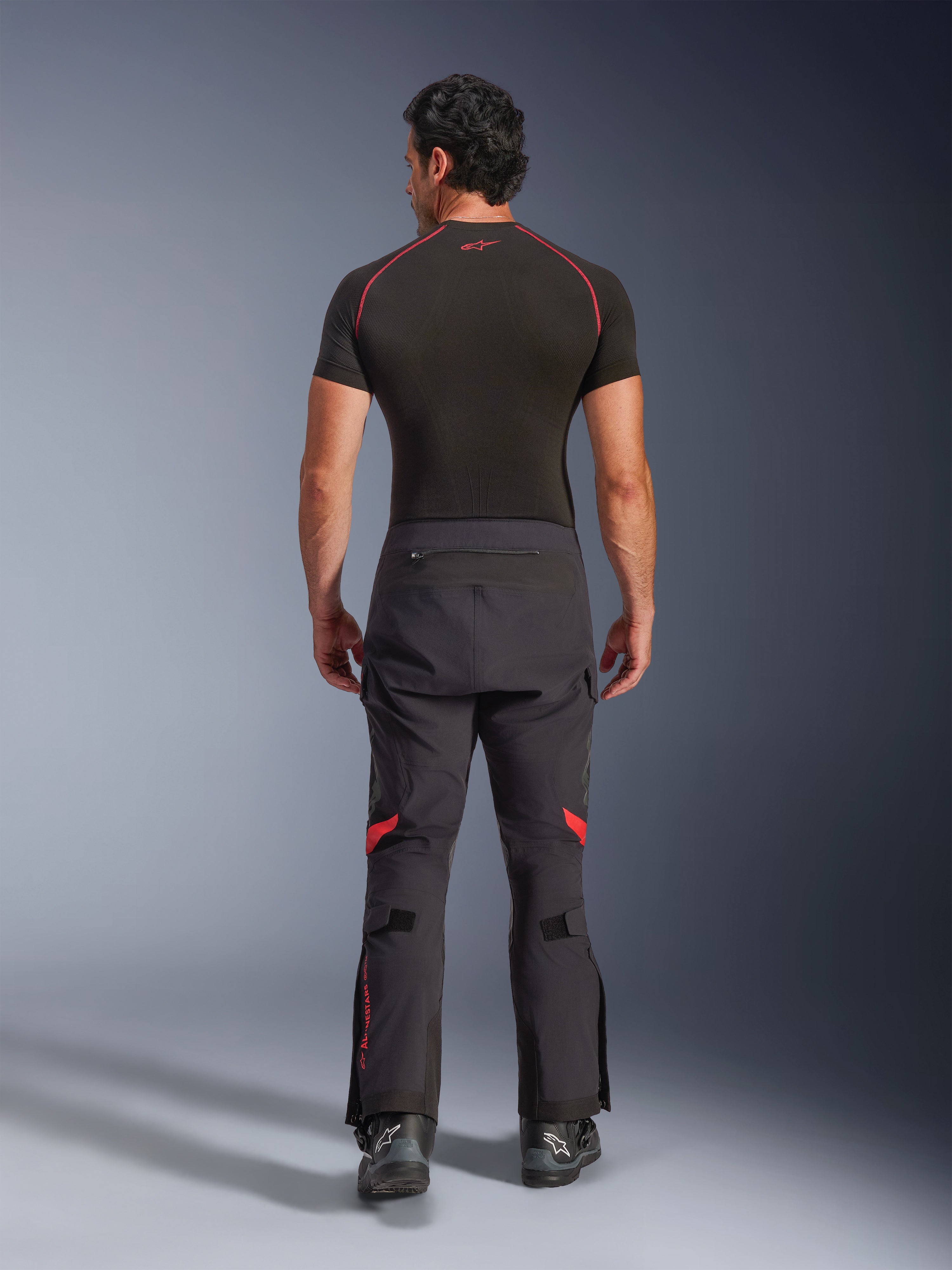 Alpinestars Halo Pro Drystar® XF laminerede bukser, adventure-motorcykelbukser, sort Dark Shadow med røde detaljer, set bagfra kombineret med et sort kortærmet basislag og sorte adventure-støvler, der viser den holdbare 3-lags laminerede konstruktion og ergonomiske pasform.