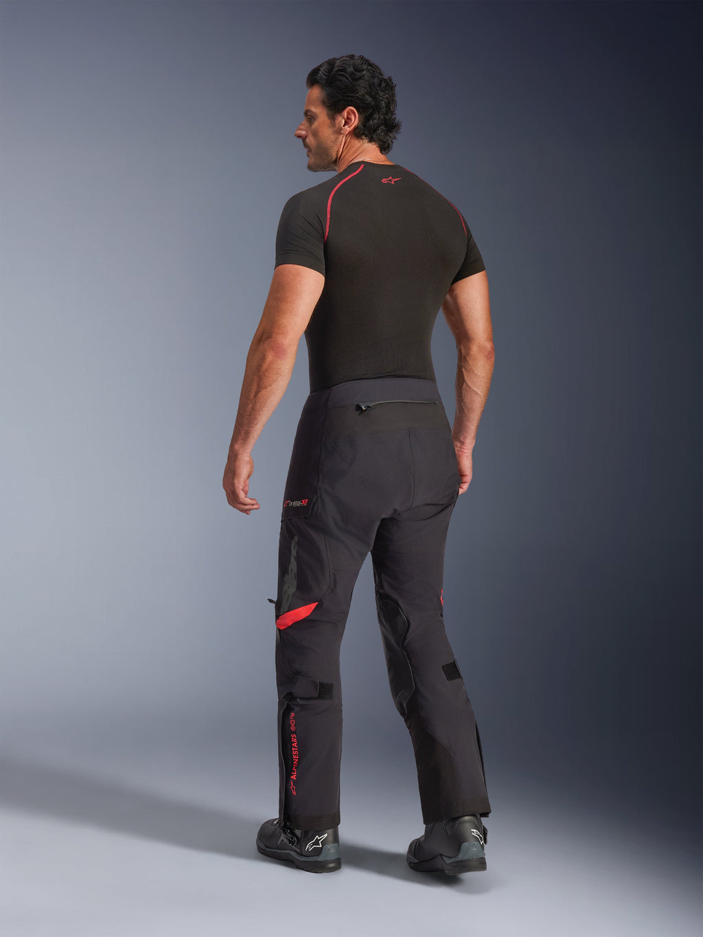 Alpinestars Halo Pro Drystar® XF laminerede bukser, Black Dark Shadow med røde detaljer, set bagfra på mandlig model, kombineret med et sort baselayer og motorcykelstøvler, teknisk adventure-touringudstyr til kørsel i alt terræn