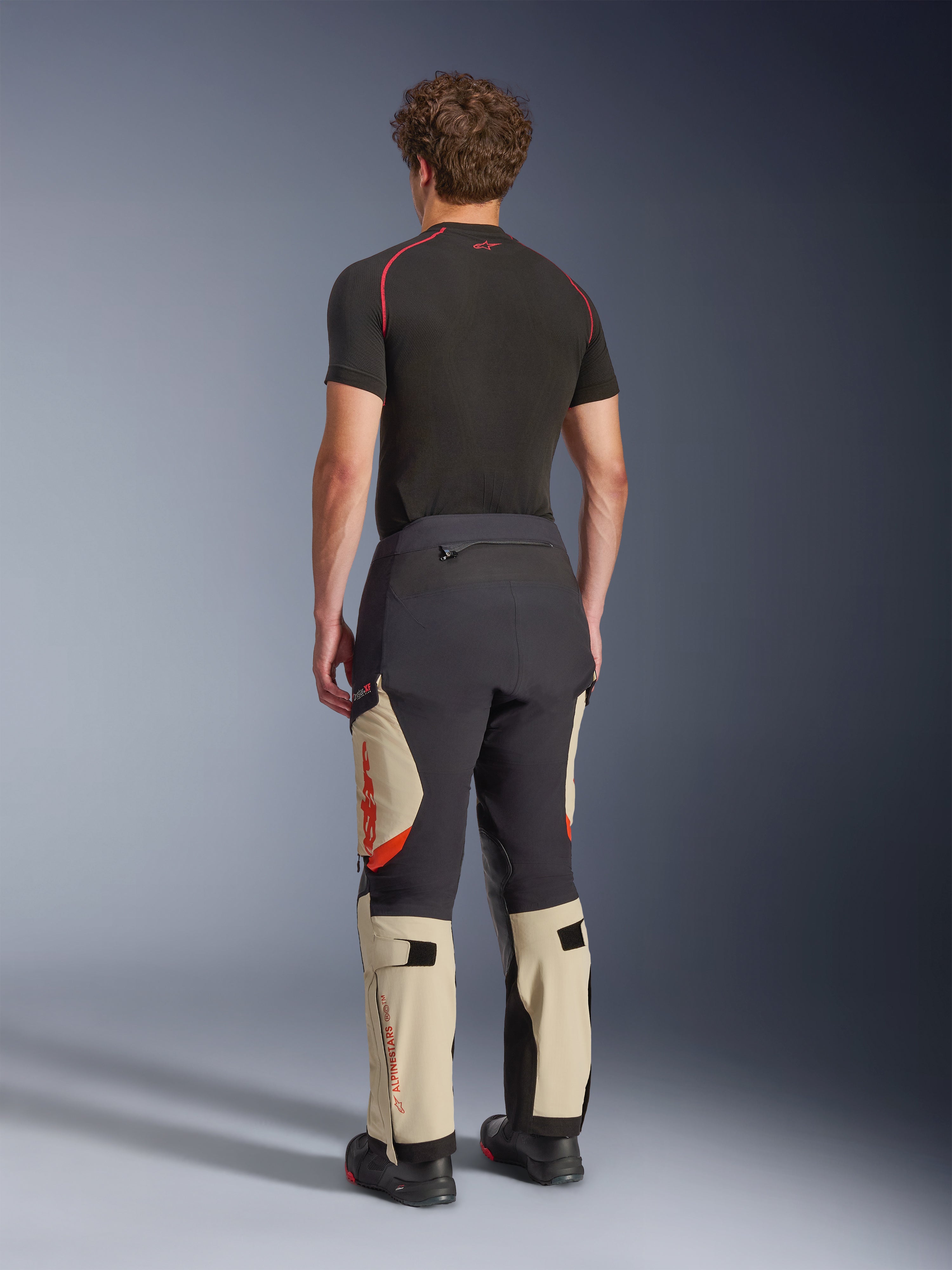 Alpinestars Halo Pro Drystar® XF laminerede bukser, adventure-motorcykelbukser, Mastic Black og Tan med orange detaljer, set bagfra viser 3-lags lamineret skalkonstruktion, kombineret med et sort basislag og adventure-støvler.