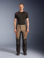 Mandlig model iført Alpinestars Andes V4 Drystar® bukser i Walnut Black, adventure motorcykelbukser kombineret med et sort baselayer og støvler, med orange lynlåsdetaljer på lårene og forstærkede sorte paneler for holdbarhed i alle terræner.