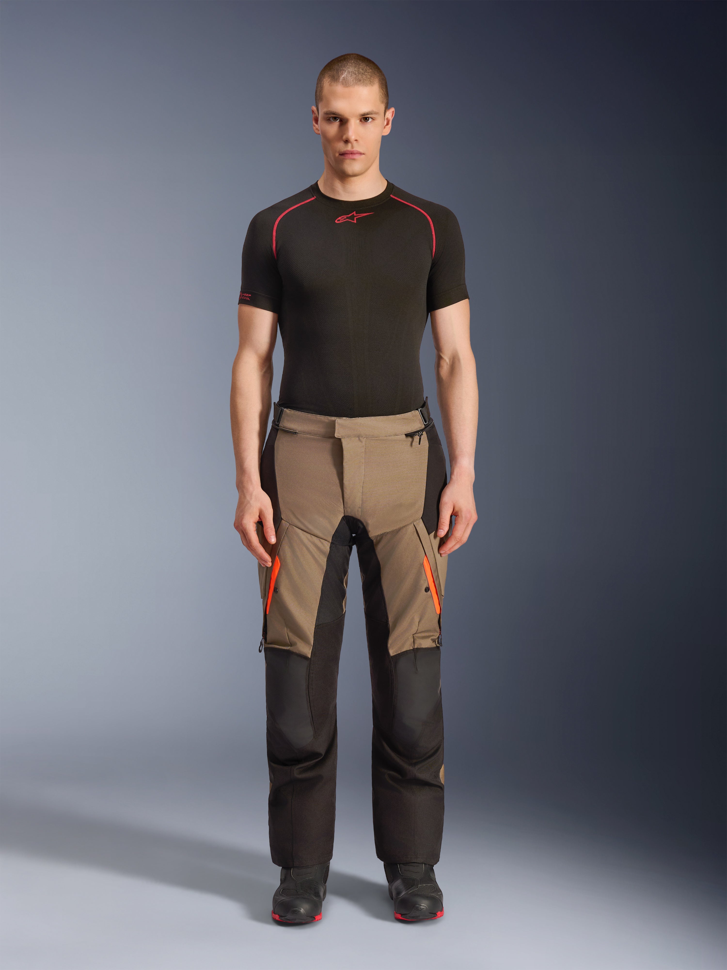 Mandlig model iført Alpinestars Andes V4 Drystar® bukser i Walnut Black, adventure motorcykelbukser kombineret med et sort baselayer og støvler, med orange lynlåsdetaljer på lårene og forstærkede sorte paneler for holdbarhed i alle terræner.