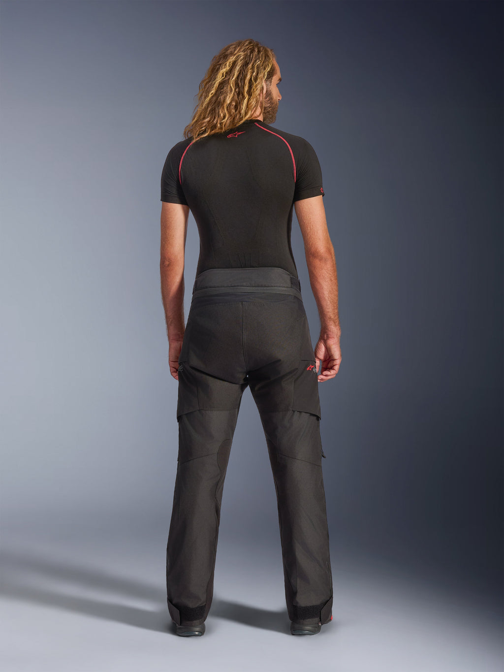 Bagfra af en model iført Alpinestars Andes V4 Drystar® bukser - Short, sorte adventure motorcykelbukser, kombineret med et sort basislag med røde syninger, der viser den slidstærke tekstilkonstruktion og ergonomiske pasform.