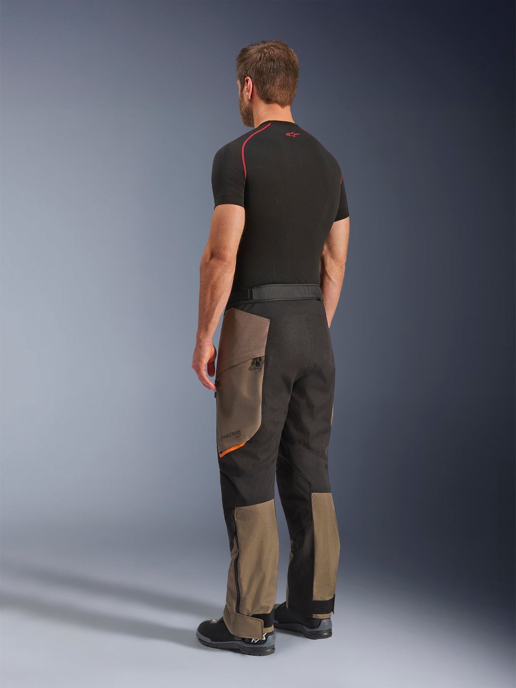 Alpinestars Cusco Drystar® bukser, adventure motorcykelbukser, Walnut Black, trekvartvisning bagfra, båret med et sort basislag og støvler, med forstærkede tekstilpaneler, cargo-lommer med lynlås på lårene og en ergonomisk touring-pasform.