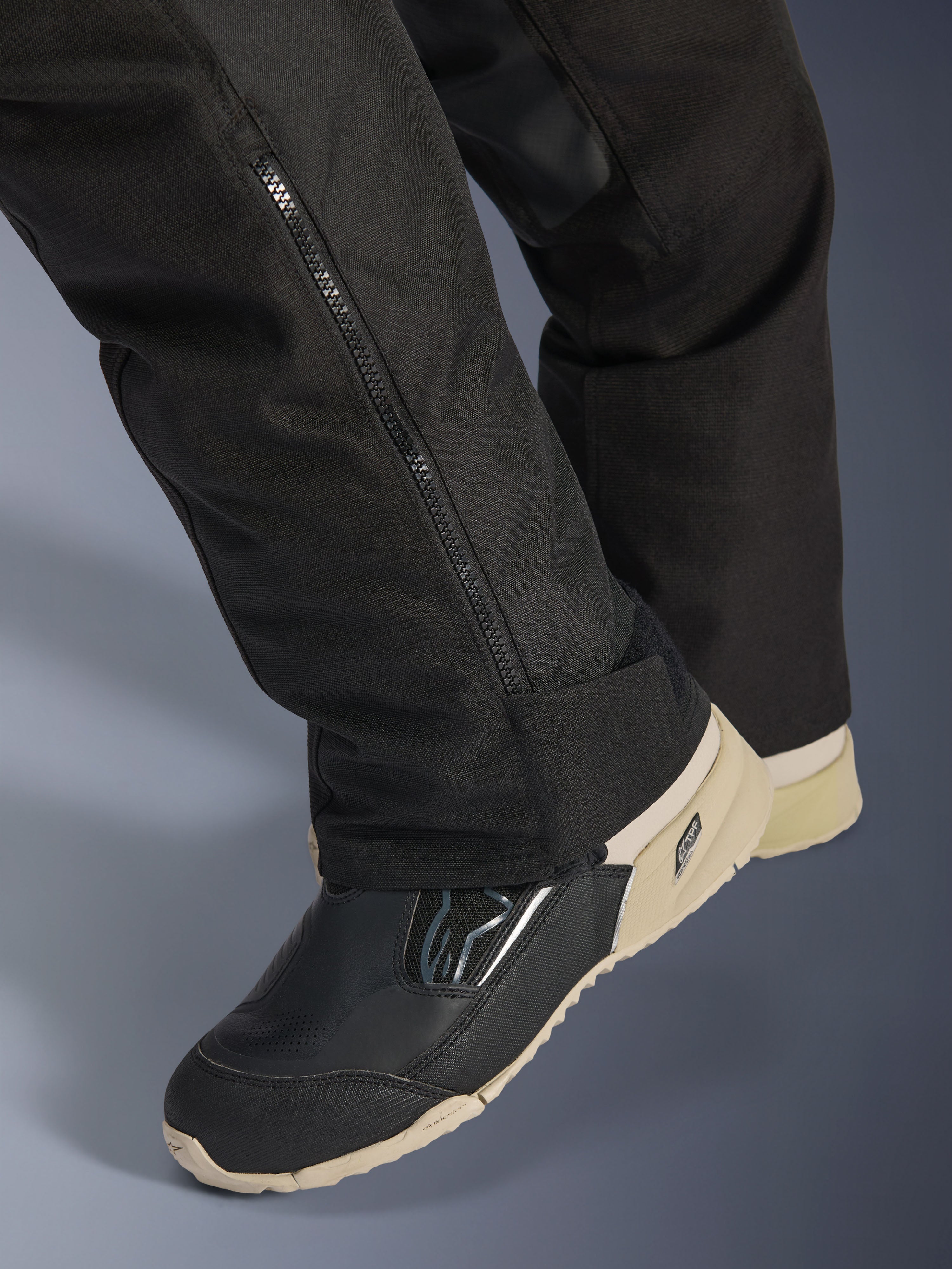 Alpinestars Stella Andes V4 Drystar® bukser, sort, nærbillede af underbenet med lynlås i siden til støvlejustering, slidstærkt tekstilmateriale, båret med sorte og sandfarvede motorcykelstøvler, adventure-klar funktionelt design