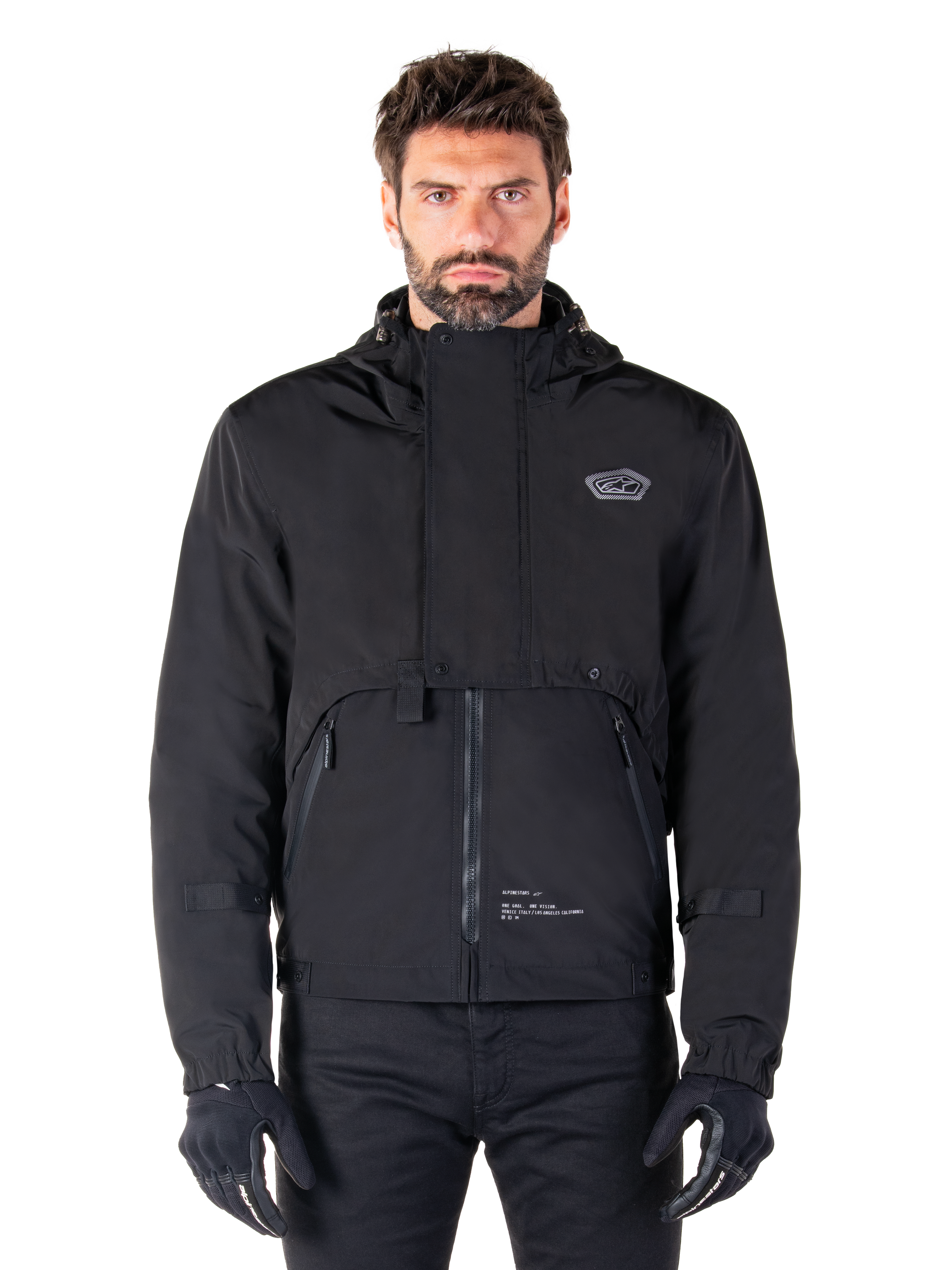 Alpinestars Mootant Waterproof Parka, sort motorcykeljakke med hætte med et modulært teknisk design, båret af en mandlig model med sorte handsker, med en unik brystklap og sidelommer med lynlås, urban og touring kørestil.