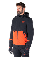 Mand iført Alpinestars Mootant Waterproof Parka i Spicy Orange og sort, en modulær motorcykeljakke med et sort øvre lag med hætte og en klar orange nedre del, stylet med sorte kørehandsker og bukser.