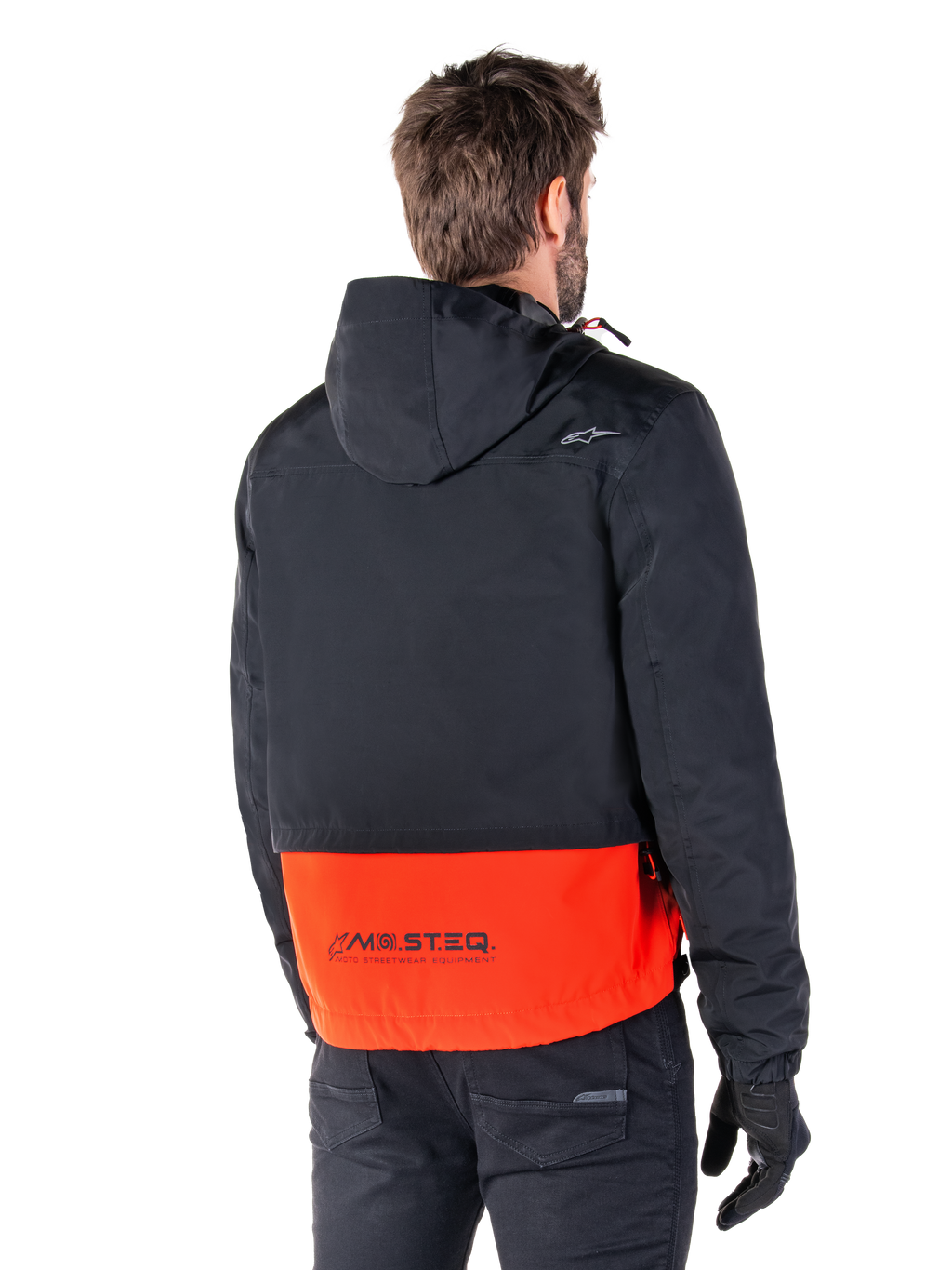 Alpinestars Mootant Vandtæt Parka, Spicy Orange og sort, set bagfra på en mandlig model, har en sort overdel med hætte og en stærk orange underdel med MO.ST.EQ.-branding, urban moderne motorcykeljakke båret med sorte bukser og handsker