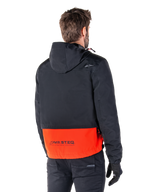 Alpinestars Mootant Vandtæt Parka, Spicy Orange og sort, set bagfra på en mandlig model, har en sort overdel med hætte og en stærk orange underdel med MO.ST.EQ.-branding, urban moderne motorcykeljakke båret med sorte bukser og handsker