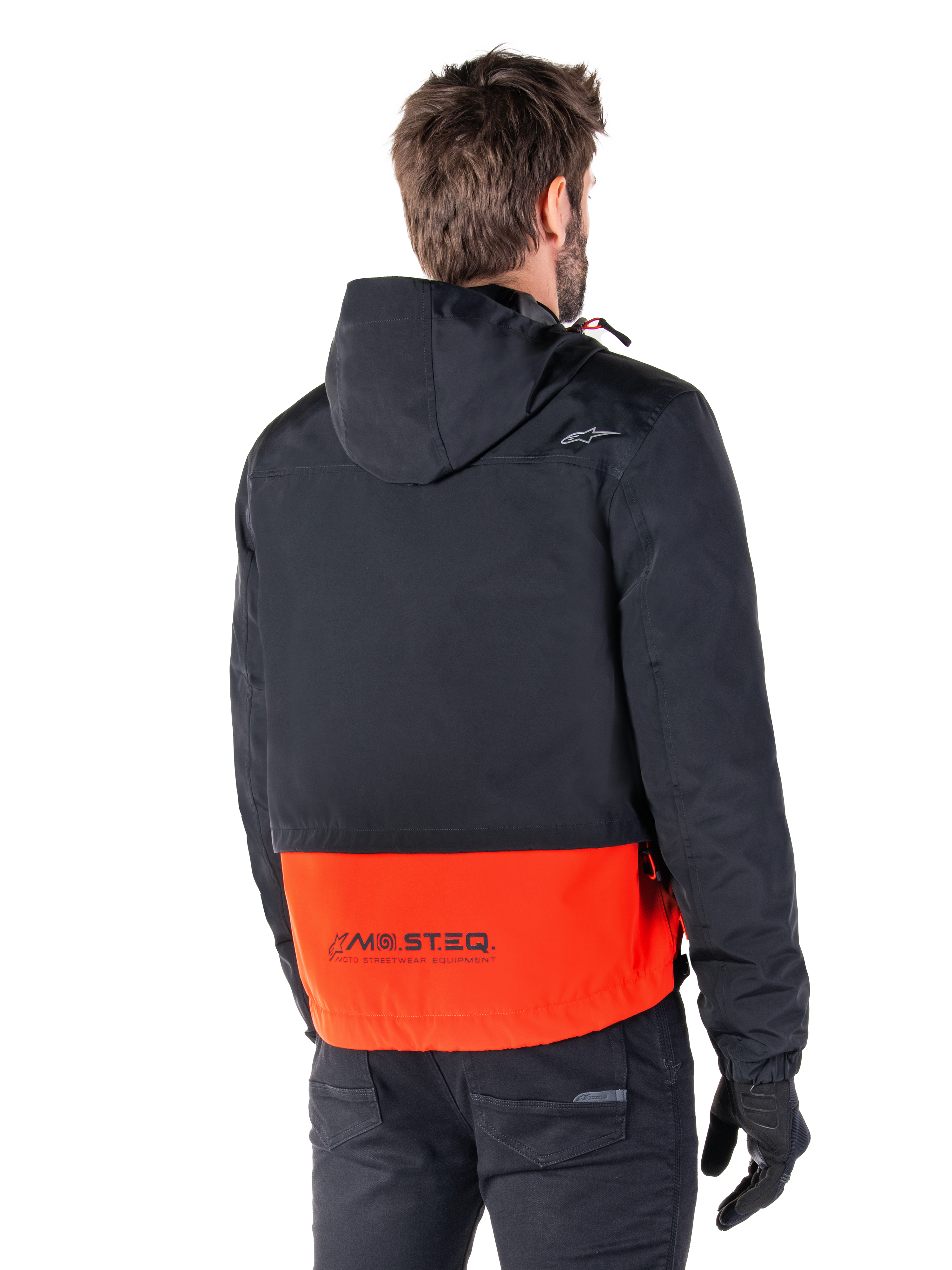 Alpinestars Mootant Vandtæt Parka, Spicy Orange og sort, set bagfra på en mandlig model, har en sort overdel med hætte og en stærk orange underdel med MO.ST.EQ.-branding, urban moderne motorcykeljakke båret med sorte bukser og handsker