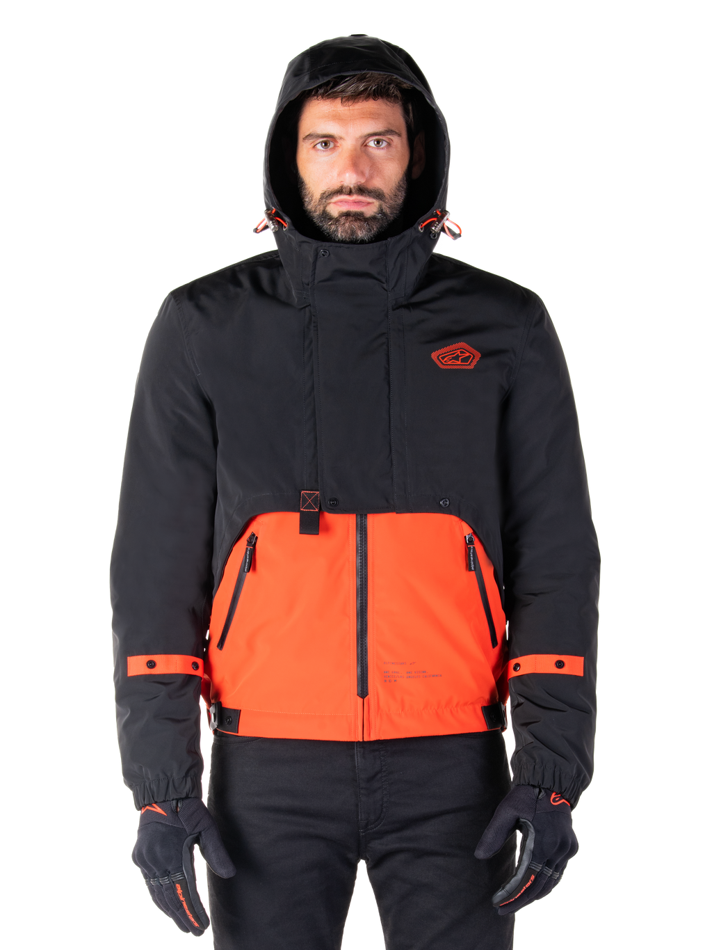 Alpinestars Mootant vandtæt parka, motorcykeljakke, Spicy Orange og sort, modulært helårsdesign med hætte, lynlåslommer foran og orange stropdetaljer på ærmerne, båret af en model med sorte handsker og bukser