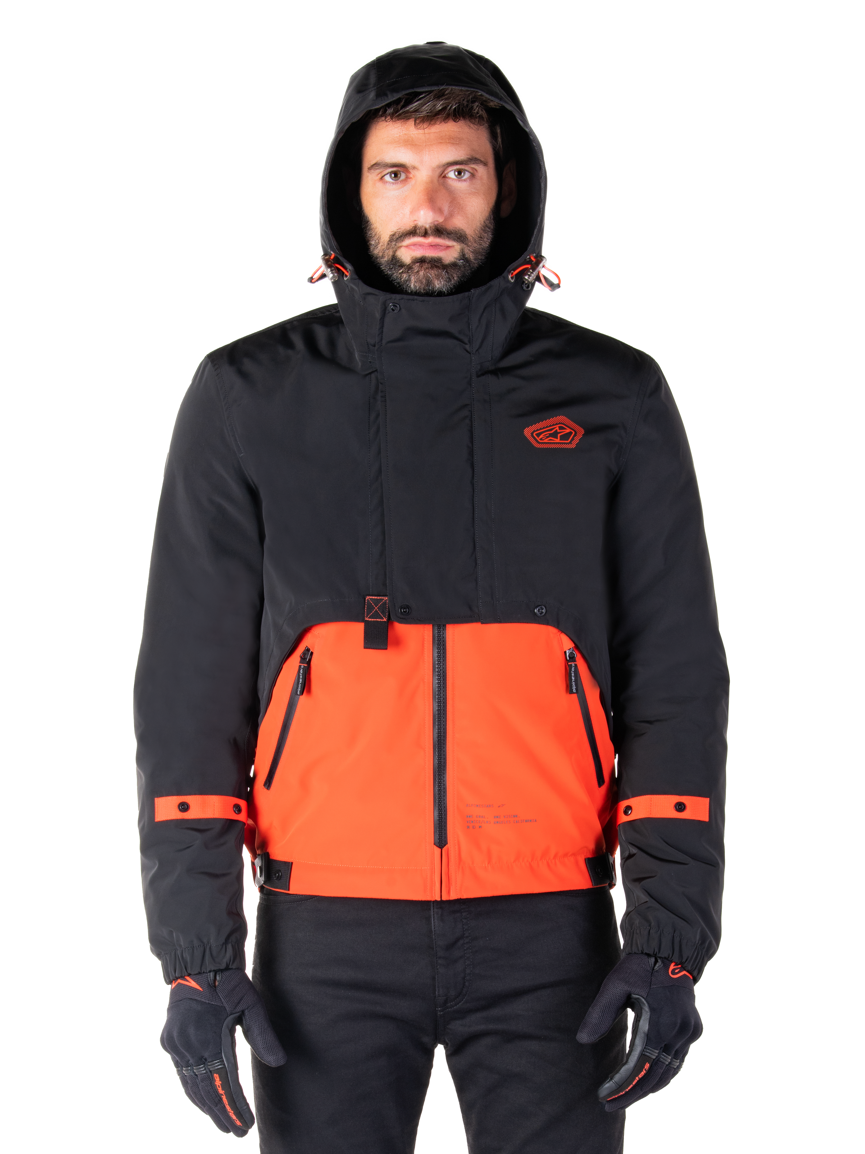 Alpinestars Mootant vandtæt parka, motorcykeljakke, Spicy Orange og sort, modulært helårsdesign med hætte, lynlåslommer foran og orange stropdetaljer på ærmerne, båret af en model med sorte handsker og bukser