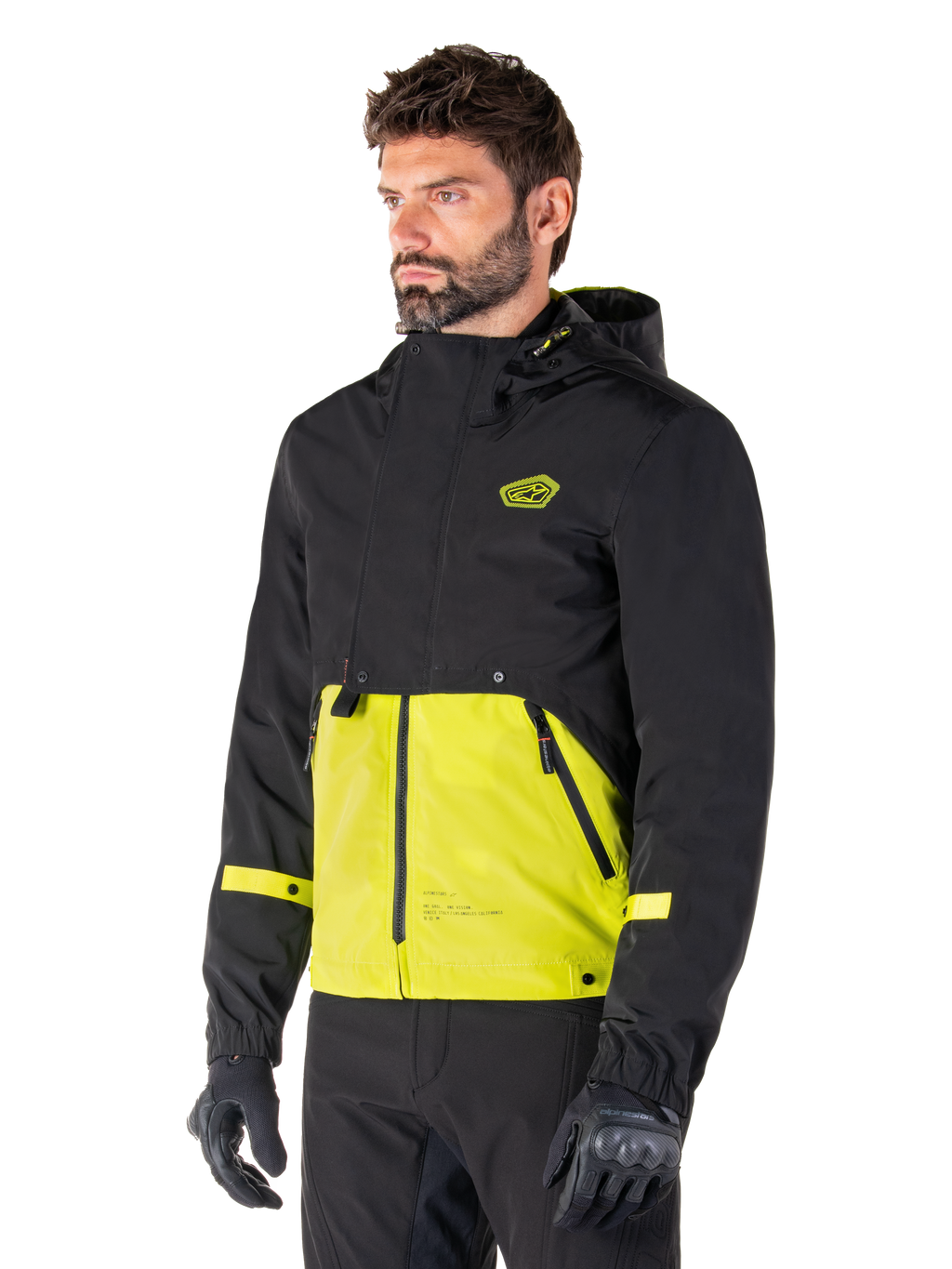 Alpinestars Mootant Waterproof Parka i Lime Punch Black, modulær motorcykeljakke båret af en mand med sorte kørehandsker og bukser, med en sort overdel og ærmer samt et markant limegrønt panel forneden, design med hætte og en utilitaristisk militærinspireret æstetik.