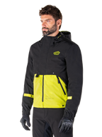 Alpinestars Mootant Waterproof Parka i Lime Punch Black, modulær motorcykeljakke båret af en mand med sorte kørehandsker og bukser, med en sort overdel og ærmer samt et markant limegrønt panel forneden, design med hætte og en utilitaristisk militærinspireret æstetik.