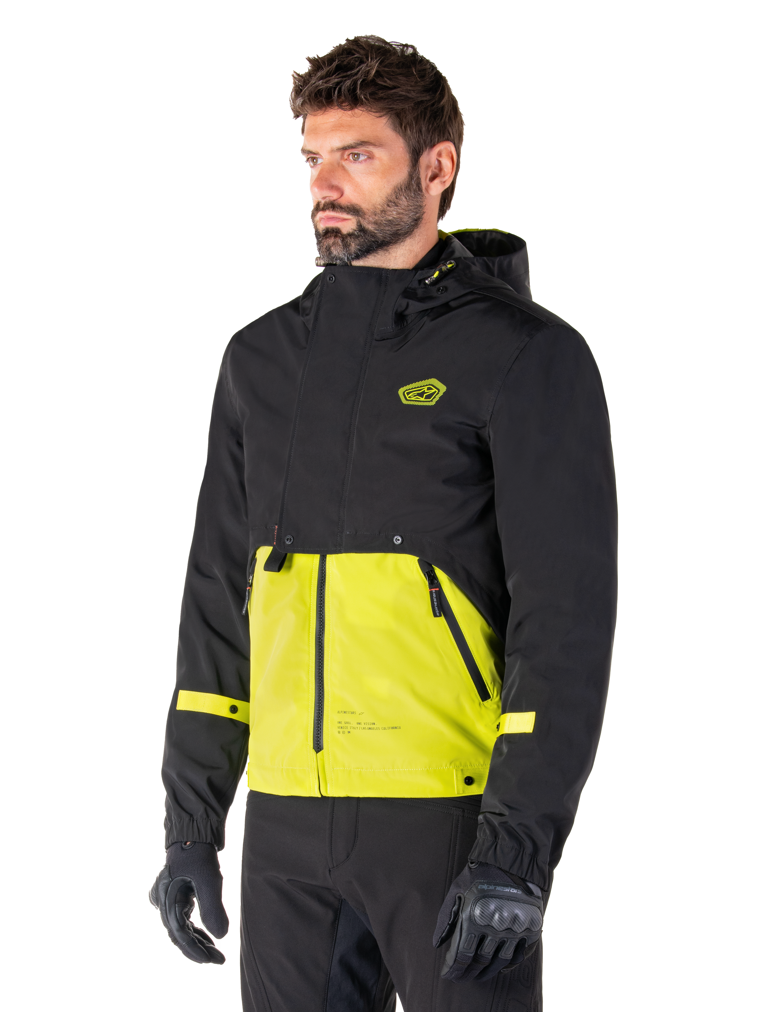 Alpinestars Mootant Waterproof Parka i Lime Punch Black, modulær motorcykeljakke båret af en mand med sorte kørehandsker og bukser, med en sort overdel og ærmer samt et markant limegrønt panel forneden, design med hætte og en utilitaristisk militærinspireret æstetik.