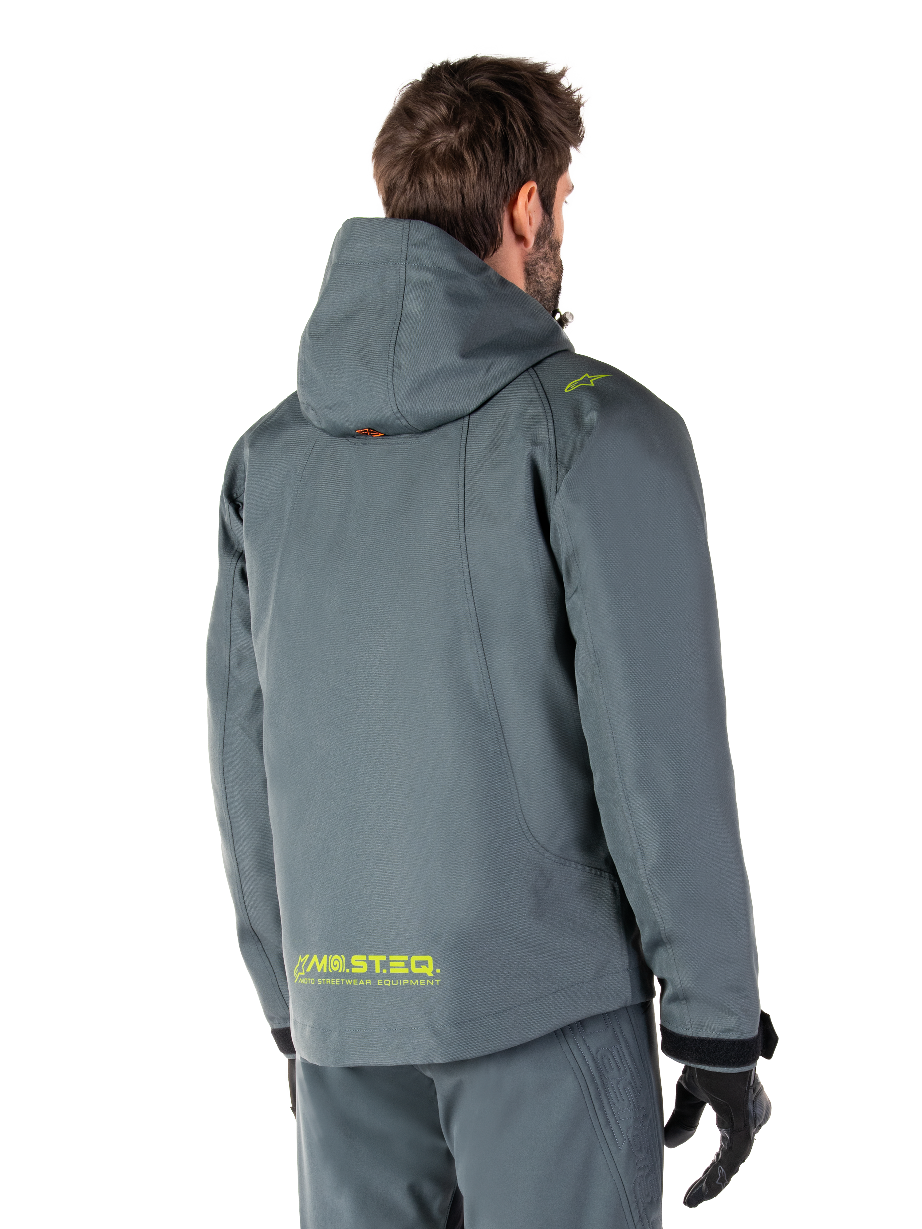 Alpinestars Morush Vandtæt Parka i Urban Gray, set bagfra, med gul MO.ST.EQ.-branding på lænden og skulderen, minimalistisk design med hætte, bæres med matchende grå kørebukser og sorte handsker, urban motorcykel-streetwear
