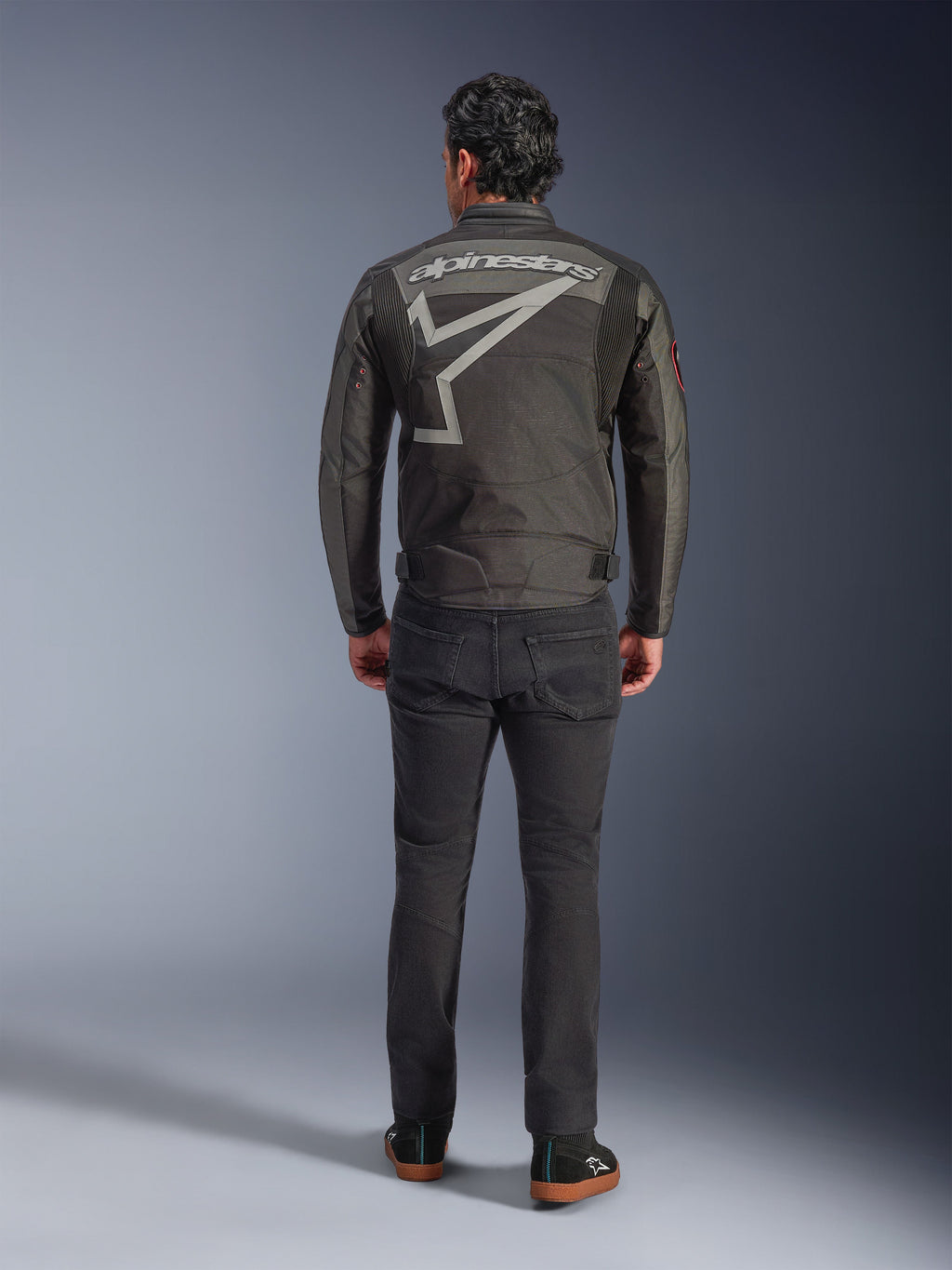 Alpinestars Axton jakke, motorcykeljakke, Black Anthracite Red Tangerine, set bagfra med stort gråt Alpinestars-logo og stjernegrafik, harmonikapaneler på skuldrene, båret med sorte bukser og afslappede sorte sko.