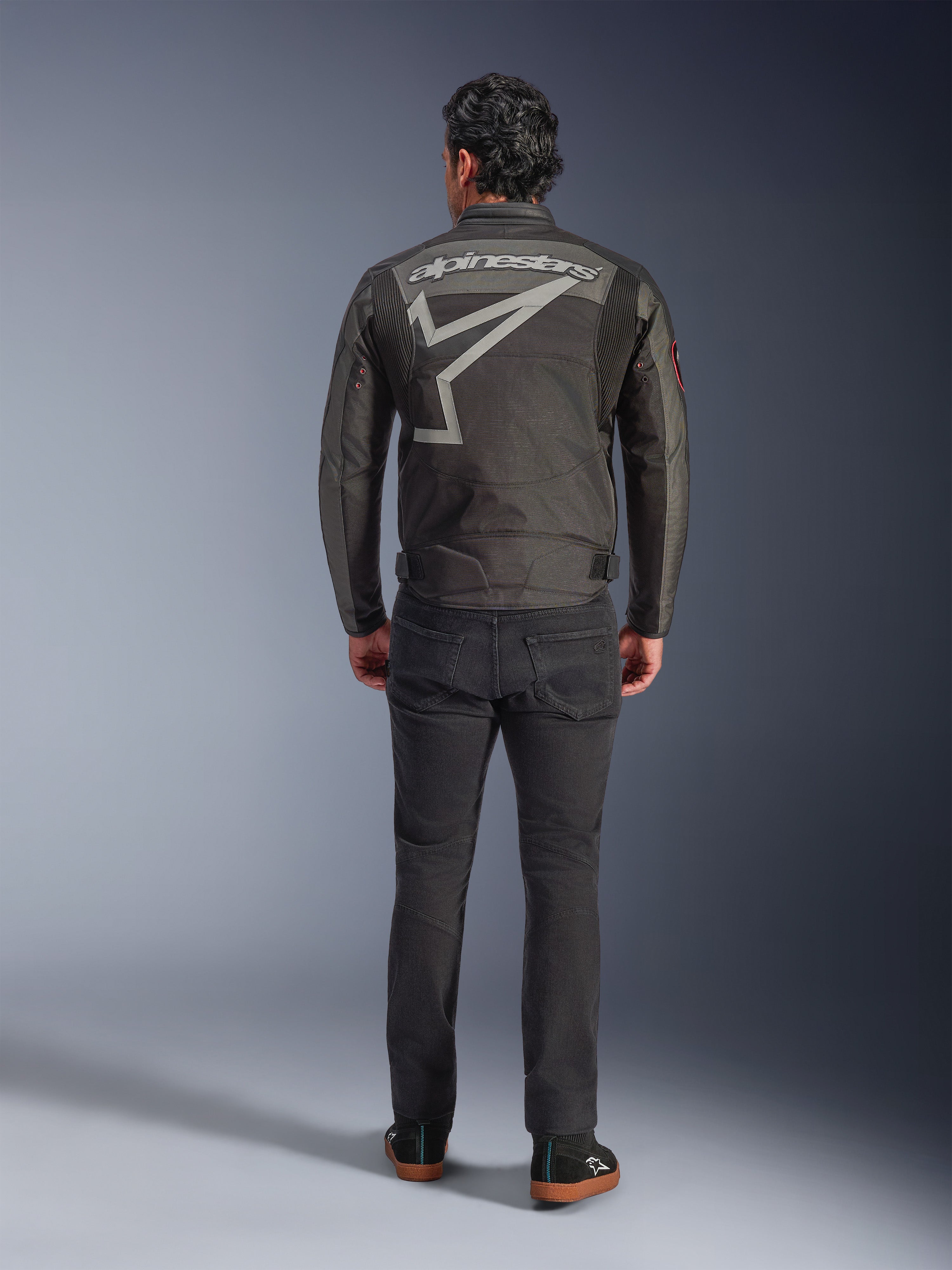 Alpinestars Axton jakke, motorcykeljakke, Black Anthracite Red Tangerine, set bagfra med stort gråt Alpinestars-logo og stjernegrafik, harmonikapaneler på skuldrene, båret med sorte bukser og afslappede sorte sko.