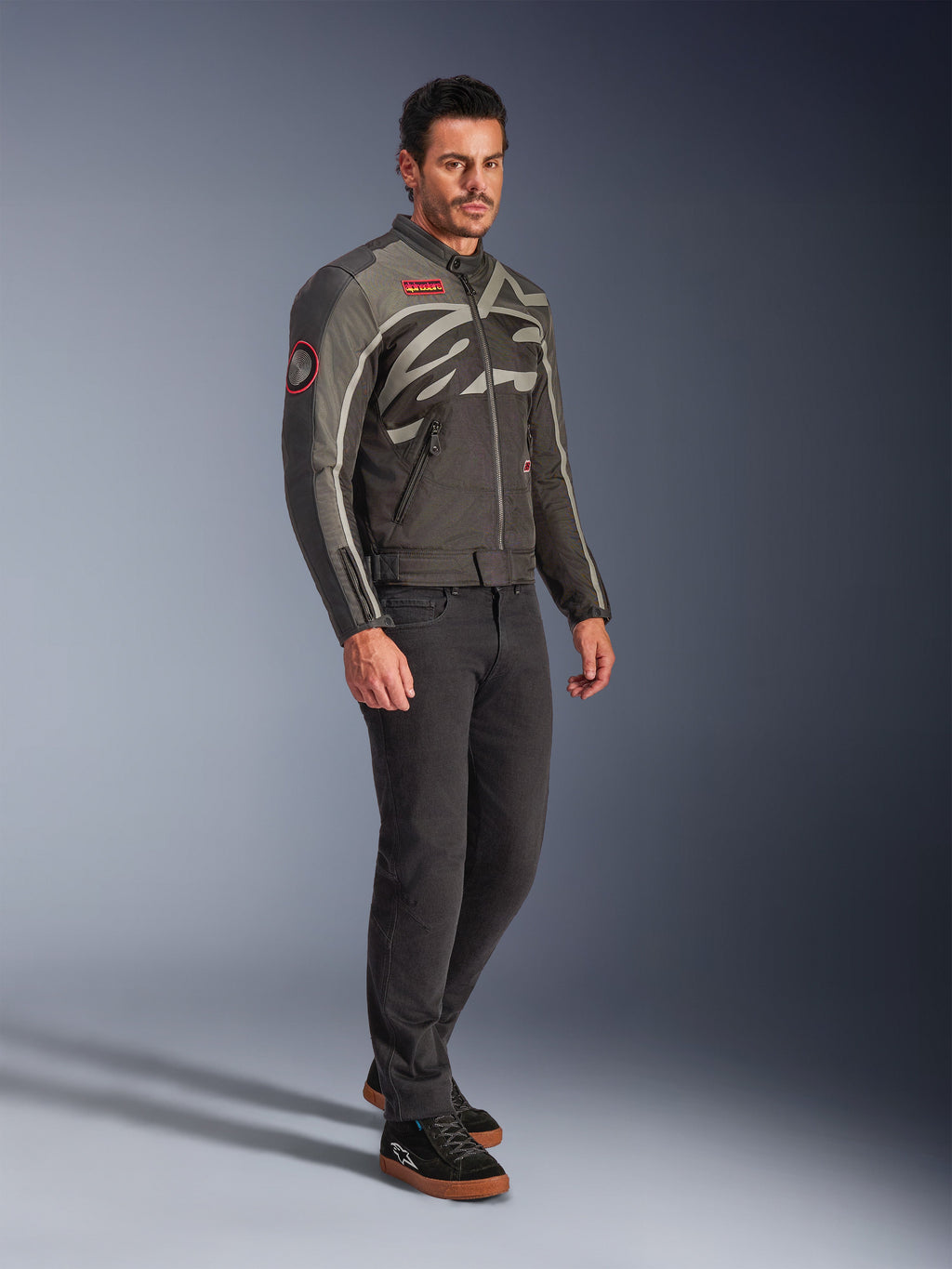 Mandlig model iført Alpinestars Axton-jakken i Black Anthracite Red Tangerine, vintage-inspireret motorcykeljakke med kropsomsluttende logo og røde detaljer, kombineret med sorte bukser og sorte køresneakers, urbant moderne motorcykeludstyr