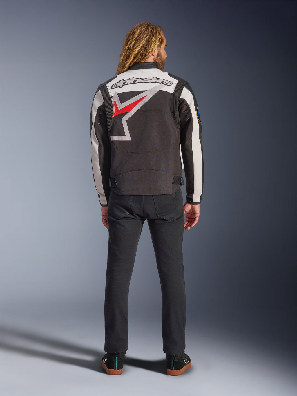 Alpinestars Axton Jacket, Motorcycle Jacket, Black Off White and Bright Red, back view on a model, featuring a large Alpinestars star logo on the back, off-white sleeve panels, worn with black pants, vintage-inspired racing teamwear design