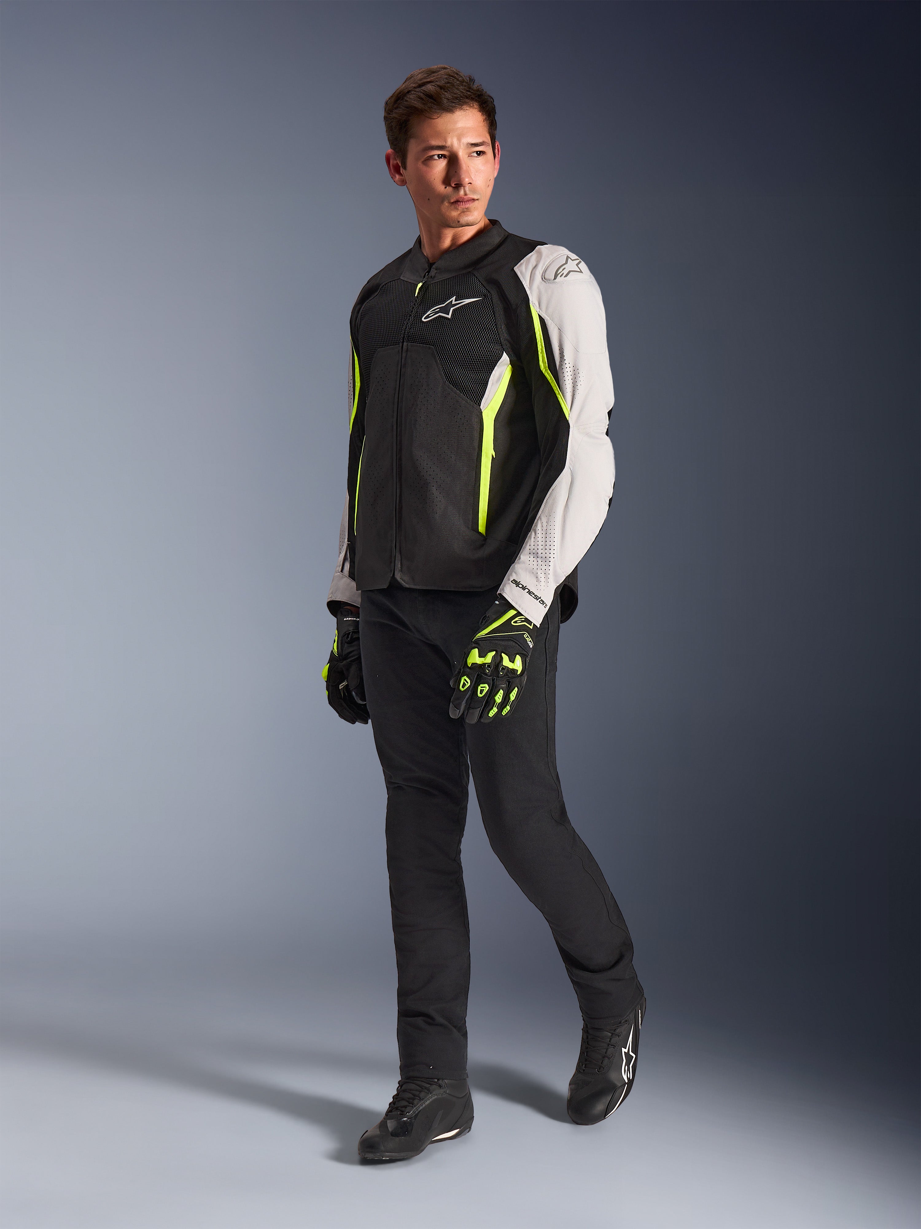 Alpinestars AST V3 Air Jacket, lightweight textile motorcycle jacket in Black, Silver, and Yellow Fluo, worn by a model with black pants and matching gloves, featuring mesh panels and laser perforations for warm-weather sport riding.