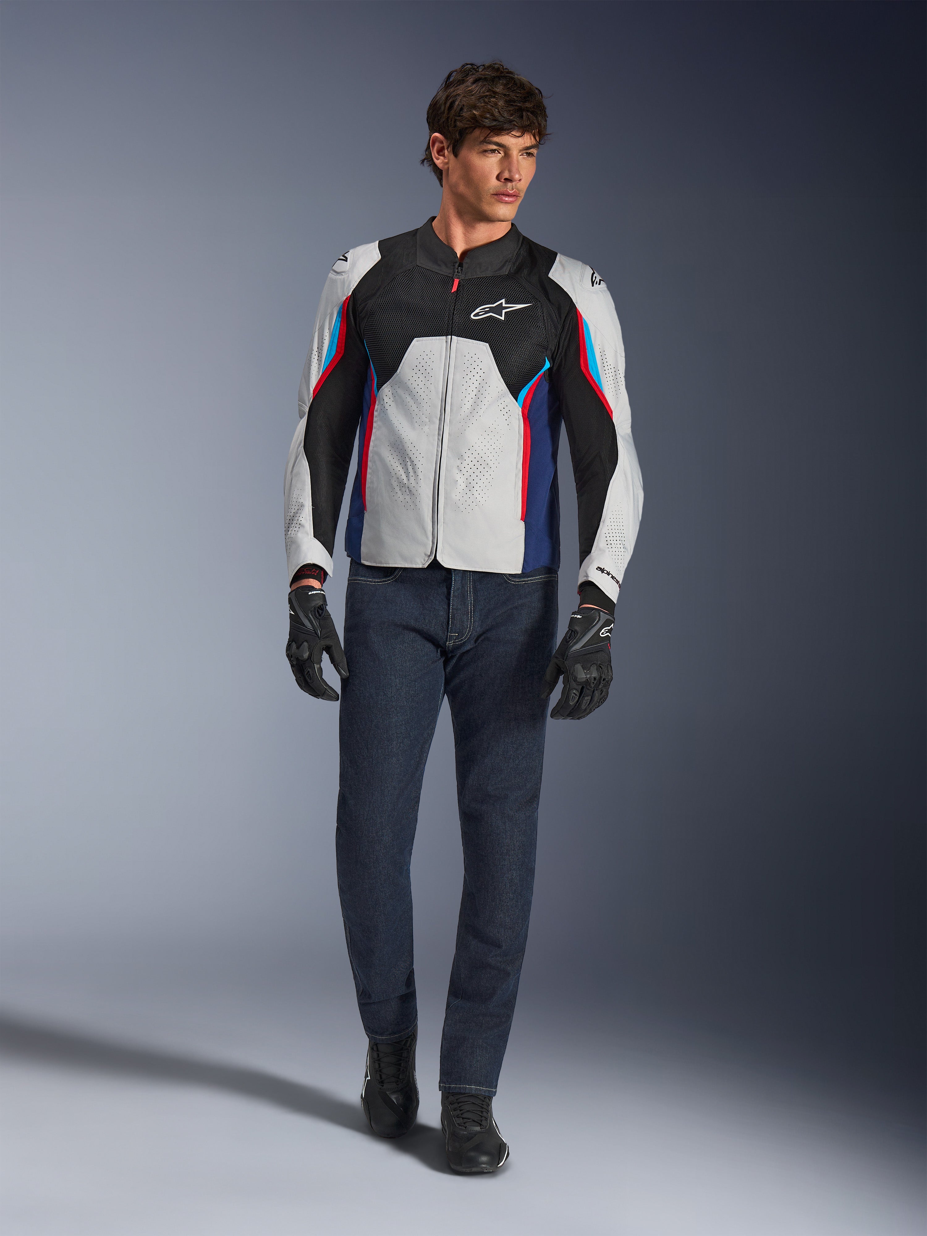 Mandlig model iført Alpinestars AST V3 Air-jakke i Silver Deep Blue Bright Red, med mesh-ventilation og perforerede paneler, kombineret med mørkeblå denim motorcykeljeans og sorte handsker, sportsoptimeret udstyr til varmt vejr.