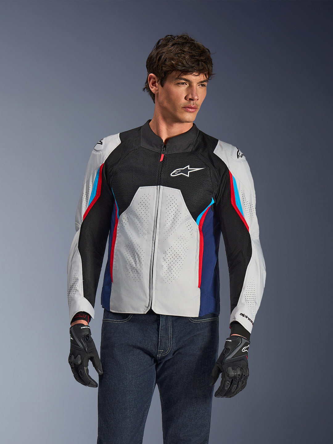 Alpinestars AST V3 Air-jakke i Silver Deep Blue Bright Red, båret af en model med mørke jeans og sorte handsker, med perforerede sølvpaneler, sort mesh-ventilation og sportslige røde og blå accenter.