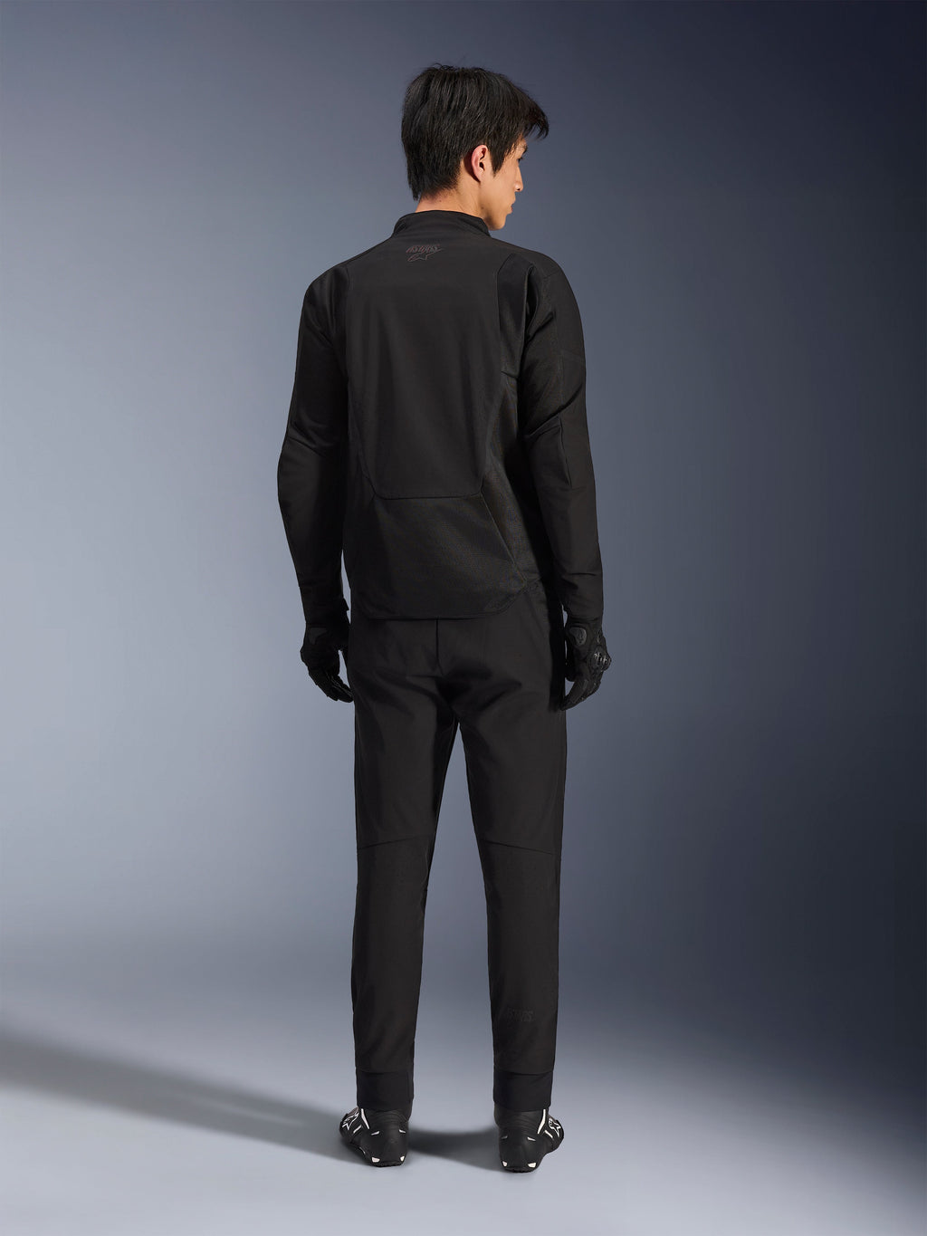 Alpinestars Aeroshell Airflow jakke i Black Anthracite, rygbillede af mandlig model i fuldt motorcykeludstyr med sorte bukser og handsker, let softshell-konstruktion med forlænget ryg for dækning
