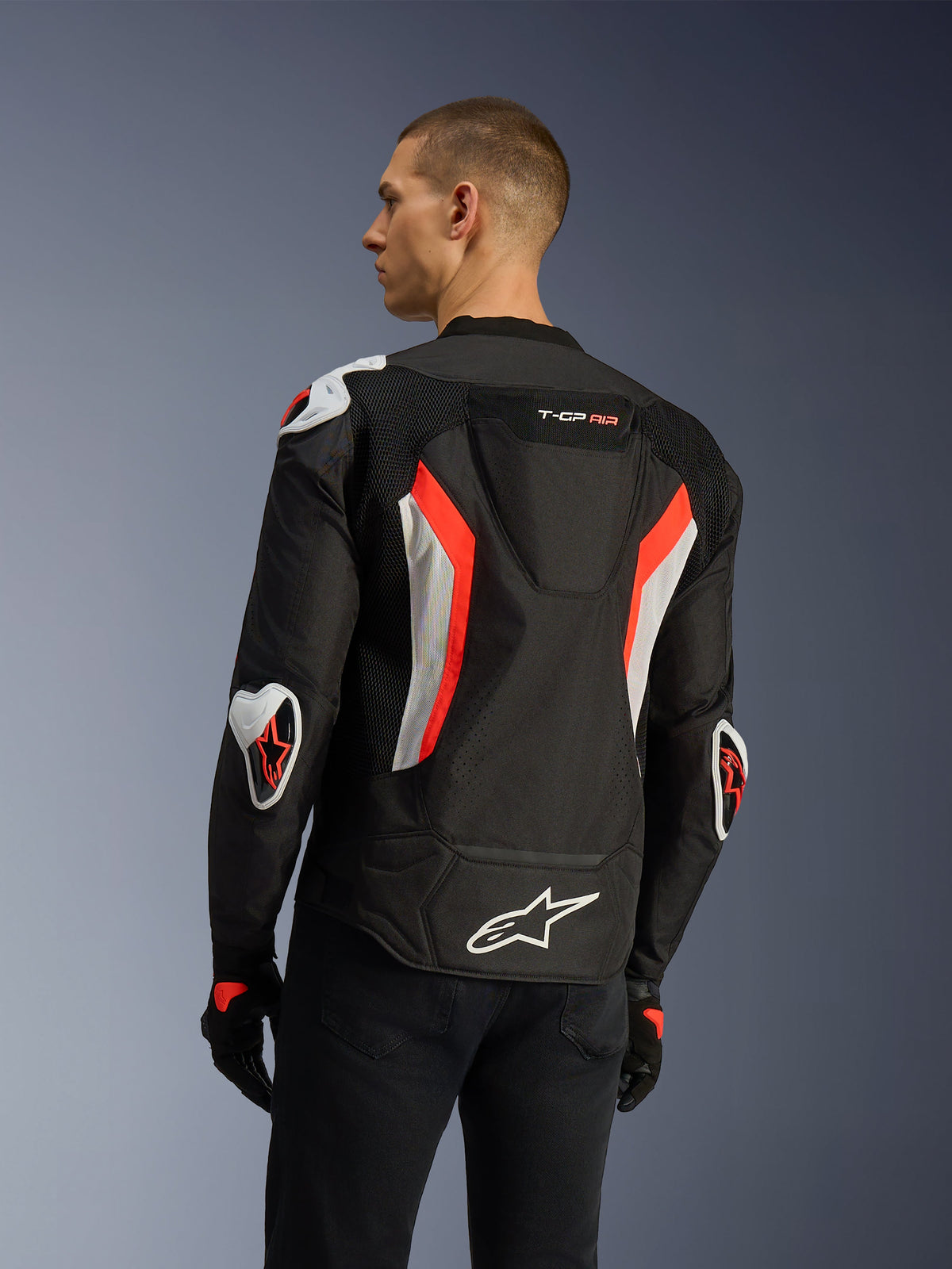 Alpinestars T-GP Air-jakke i Black Red Fluo, set bagfra i trekvart-profil på en model, der fremhæver den aerodynamiske pukkel, Racing GP DFS ekstern beskyttelse på skuldre og albuer, laserperforerede mesh-paneler, hvide og røde fluo-detaljer, sport-cut motorcykeljakke i tekstil.