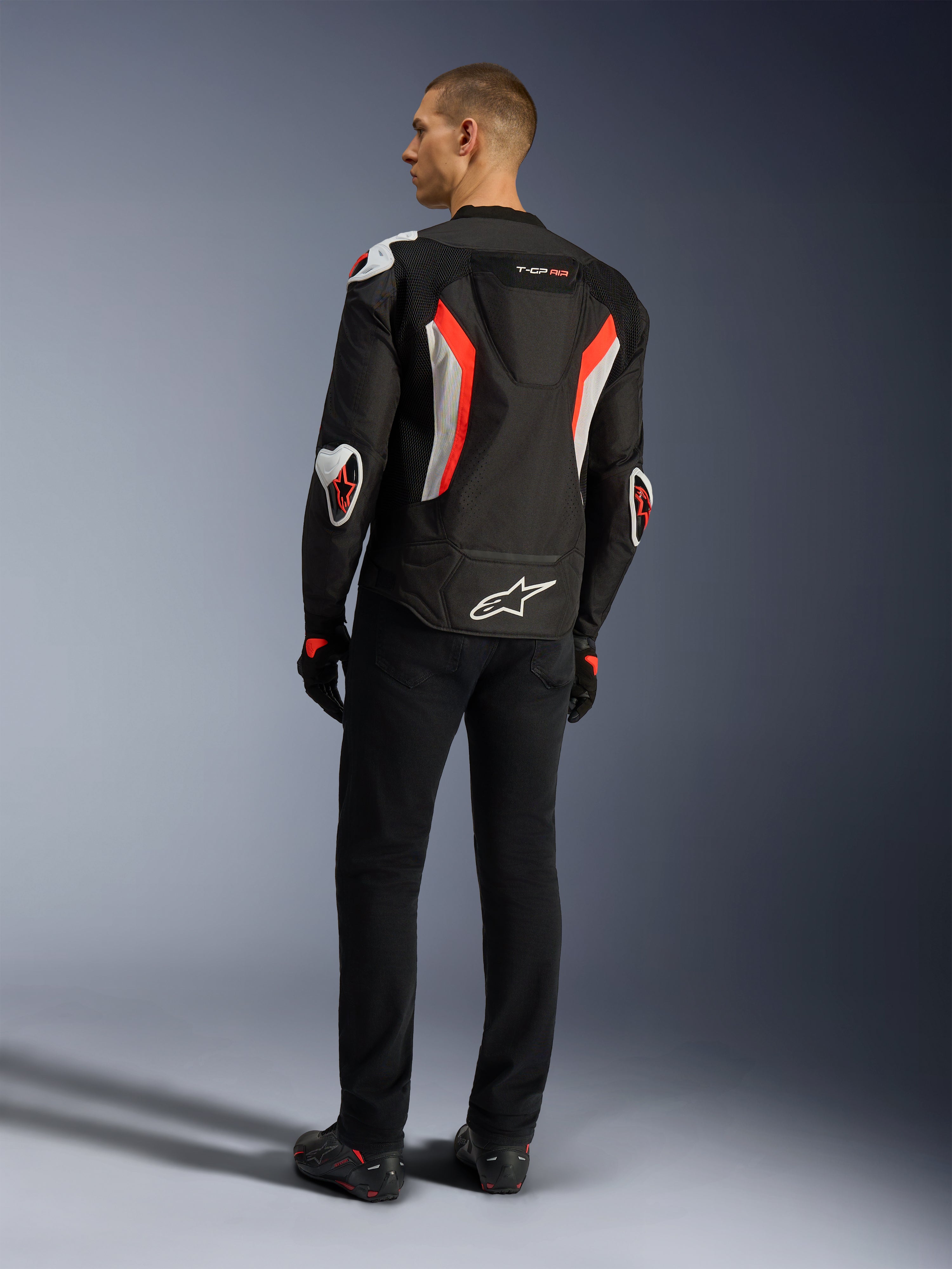 Alpinestars T-GP Air-jakke, Black Red Fluo motorcykeljakke, set bagfra på en model, med ventilationspaneler i mesh, aerodynamisk pukkel og hvide udvendige beskyttere på albuer og skuldre, båret med sorte bukser og køresko