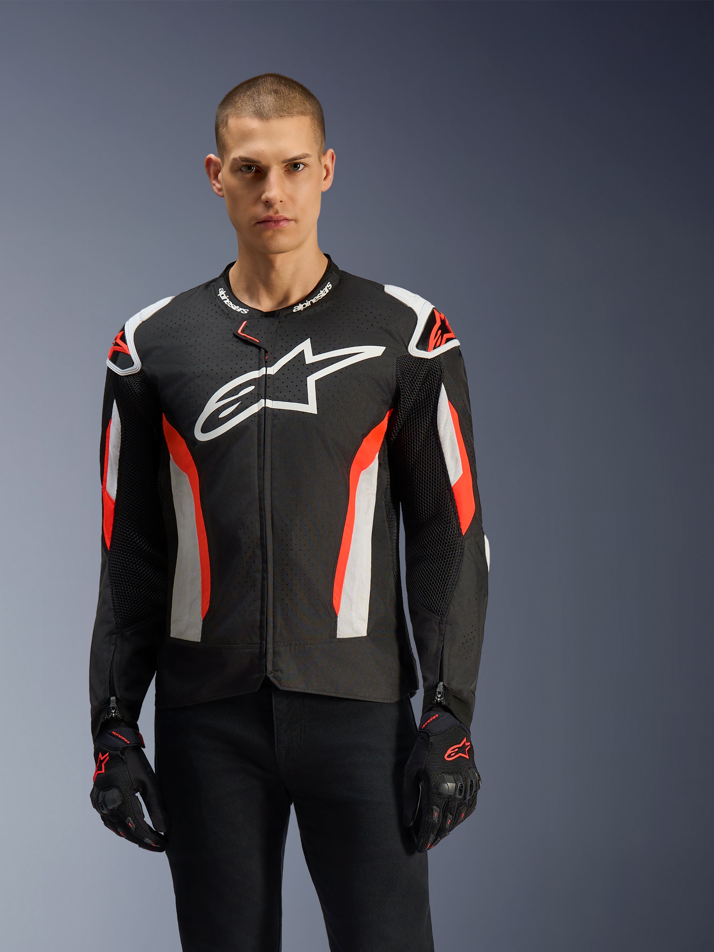 Mandlig model iført Alpinestars T-GP Air-jakke, sort med rød fluo og hvide detaljer, stort Alpinestars-logo på brystet, med laserperforerede paneler og skulderbeskyttelse, båret med sorte kørehandsker, sportsmotorcykeludstyr til varmt vejr