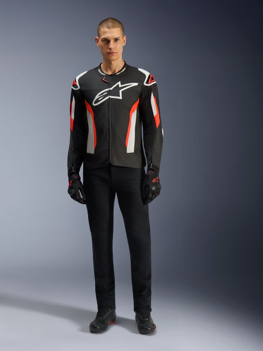 Mandlig model iført Alpinestars T-GP Air-jakke i Black Red Fluo, tekstil motorcykeljakke med hvide og klare røde detaljer, stort hvidt Alpinestars-logo på brystet, med mesh-ventilationspaneler, kombineret med sorte tekniske bukser og kørehandsker, sportsskåret udstyr