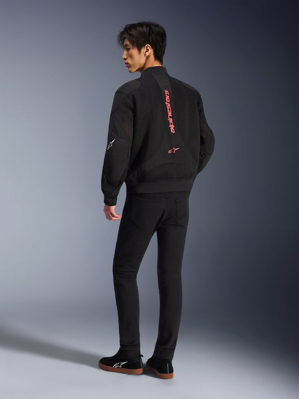 Mandlig model iført Alpinestars Flight Air-jakke i Black Bright Red, set bagfra viser lodret rød branding på ryggen og 3D-mesh-konstruktion, kombineret med sorte bukser og sneakers, urban motorcykelstil