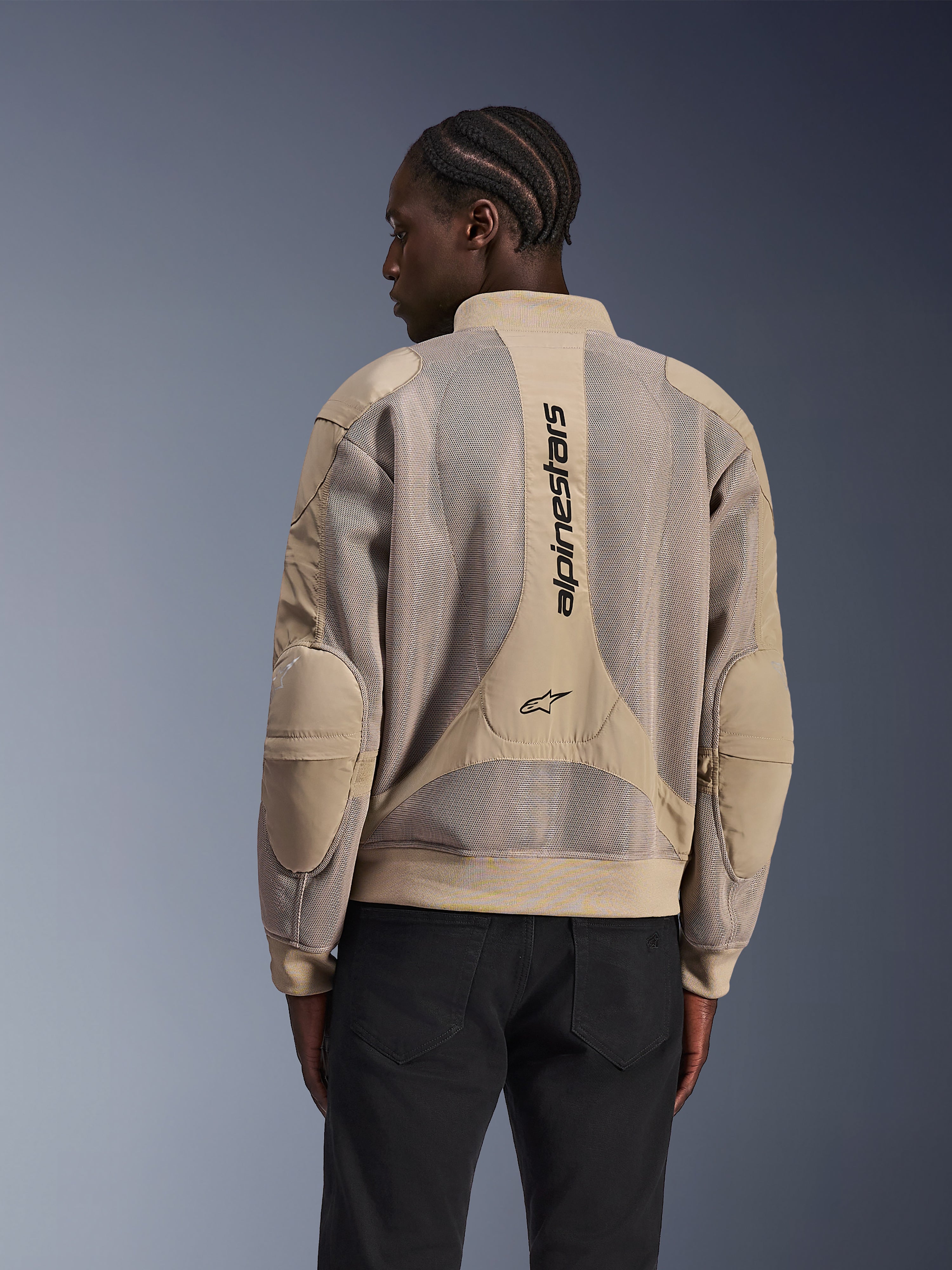 Alpinestars Flight Air-jakke, urban motorcykeljakke, tan, set bagfra med udvidede 3D-mesh-paneler og lodret Alpinestars-branding på ryggen, bomber-inspireret design med ribkant, båret af model i sorte bukser