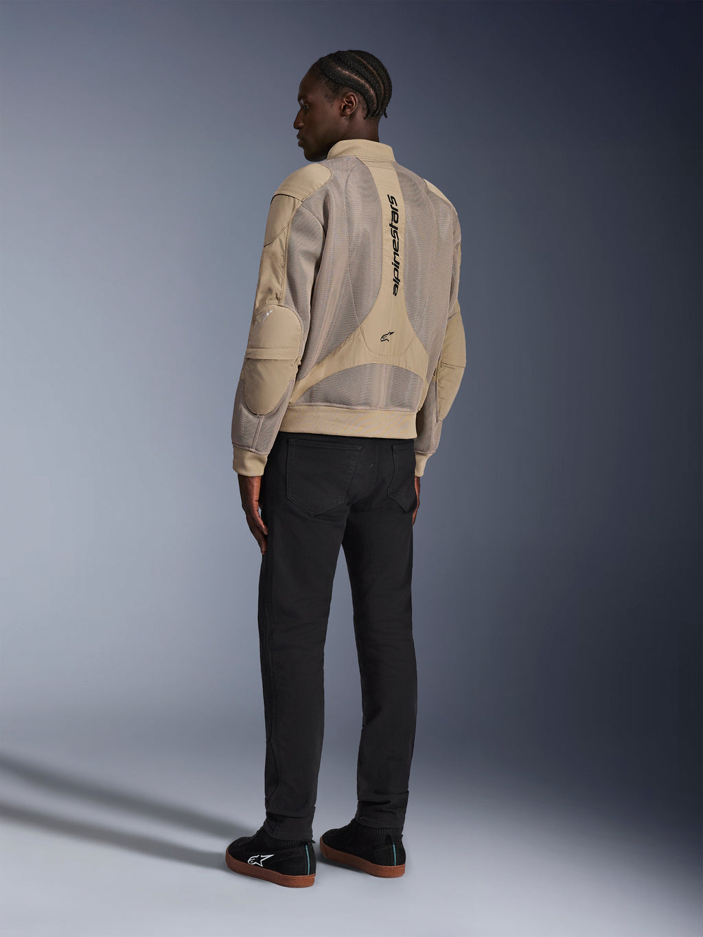 Alpinestars Flight Air-jakke, urban motorcykeljakke i tekstil, brun, set bagfra med store 3D-mesh-paneler og lodret logo, bomber-silhuet med ribstrikket krave og kant, kombineret med sorte bukser og sorte sko for en street-inspireret kørestil.