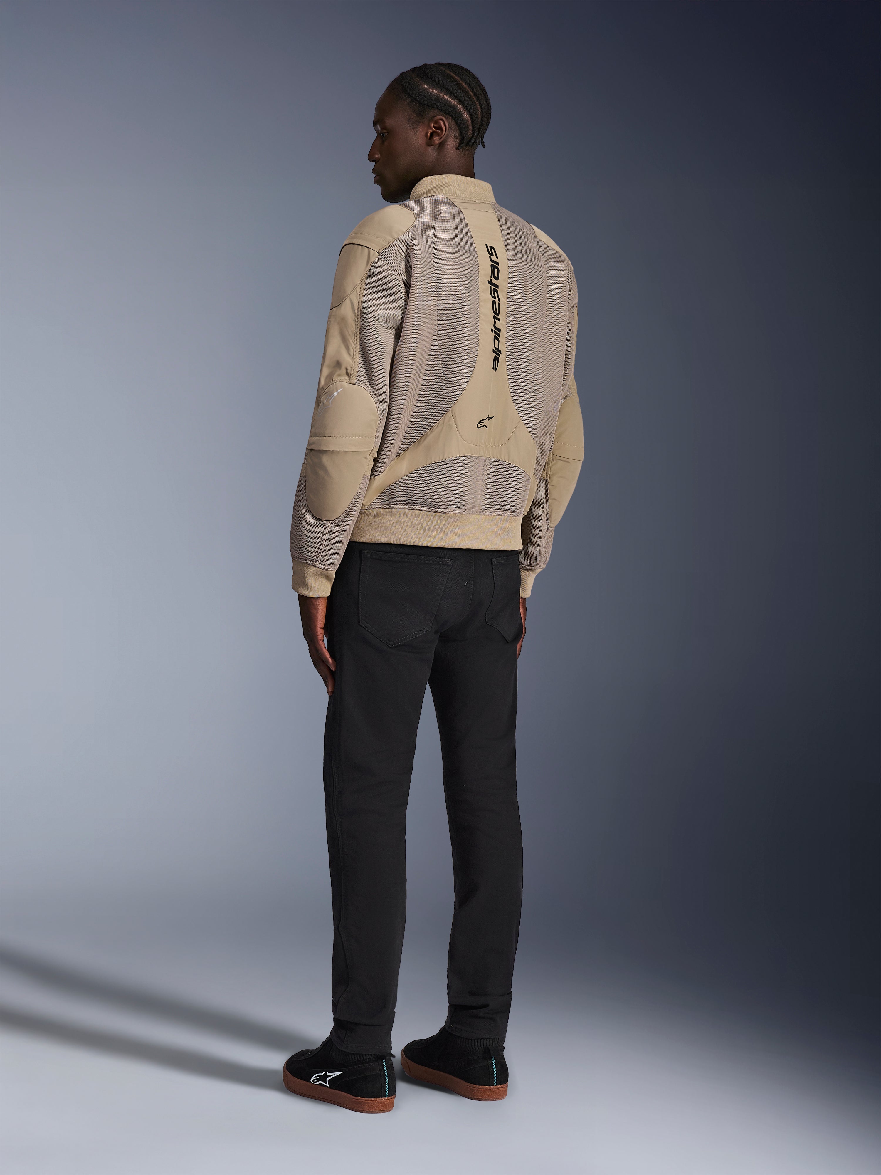 Alpinestars Flight Air-jakke, urban motorcykeljakke i tekstil, brun, set bagfra med store 3D-mesh-paneler og lodret logo, bomber-silhuet med ribstrikket krave og kant, kombineret med sorte bukser og sorte sko for en street-inspireret kørestil.