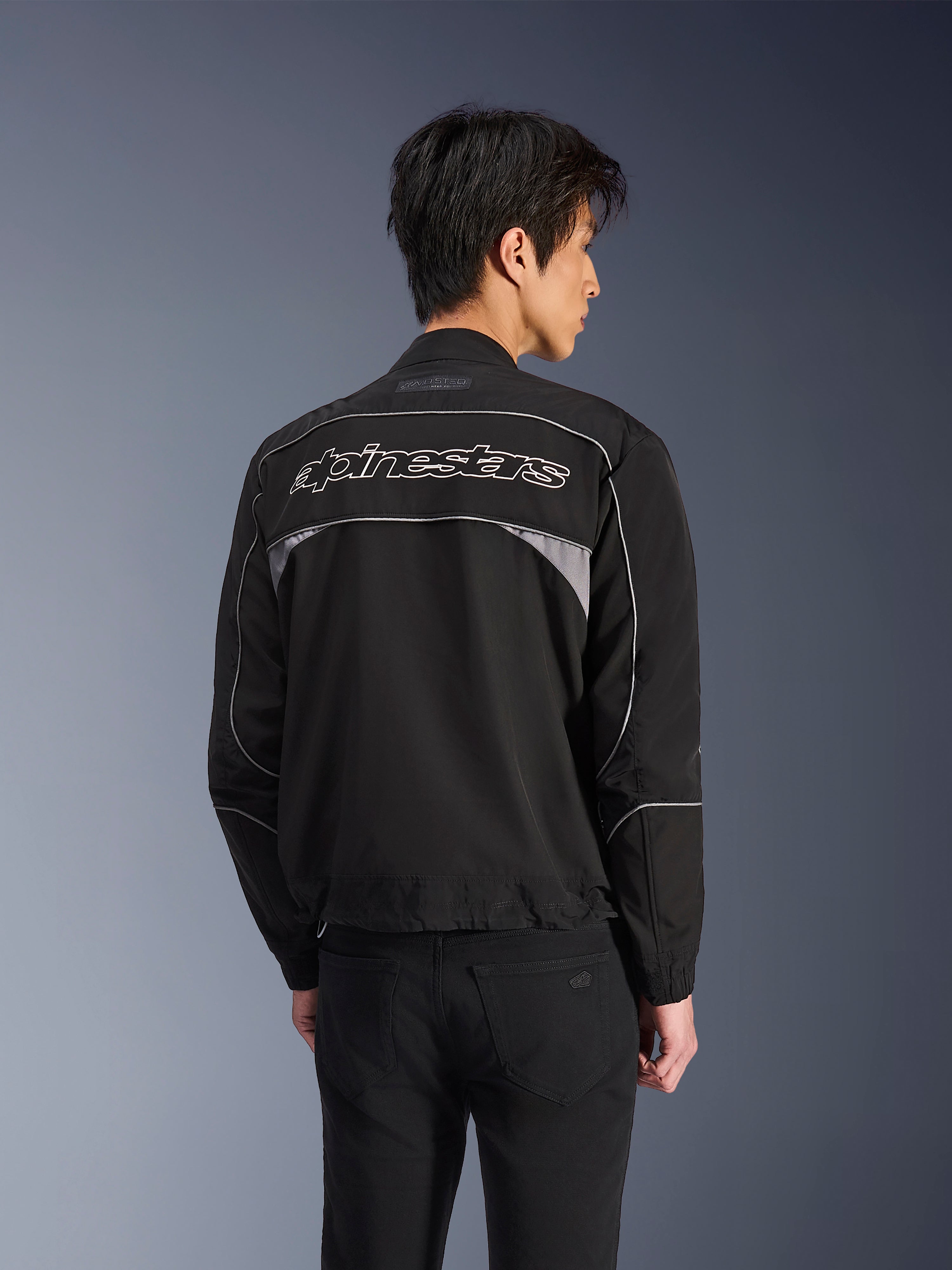 Alpinestars Unite jakke, urban motorcykeljakke, sort med mørkegrå detaljer, rygvisning der viser hvidt Alpinestars-navnetræk hen over skuldrene, reflekterende piping og paneler, minimalistisk softshell streetwear-design